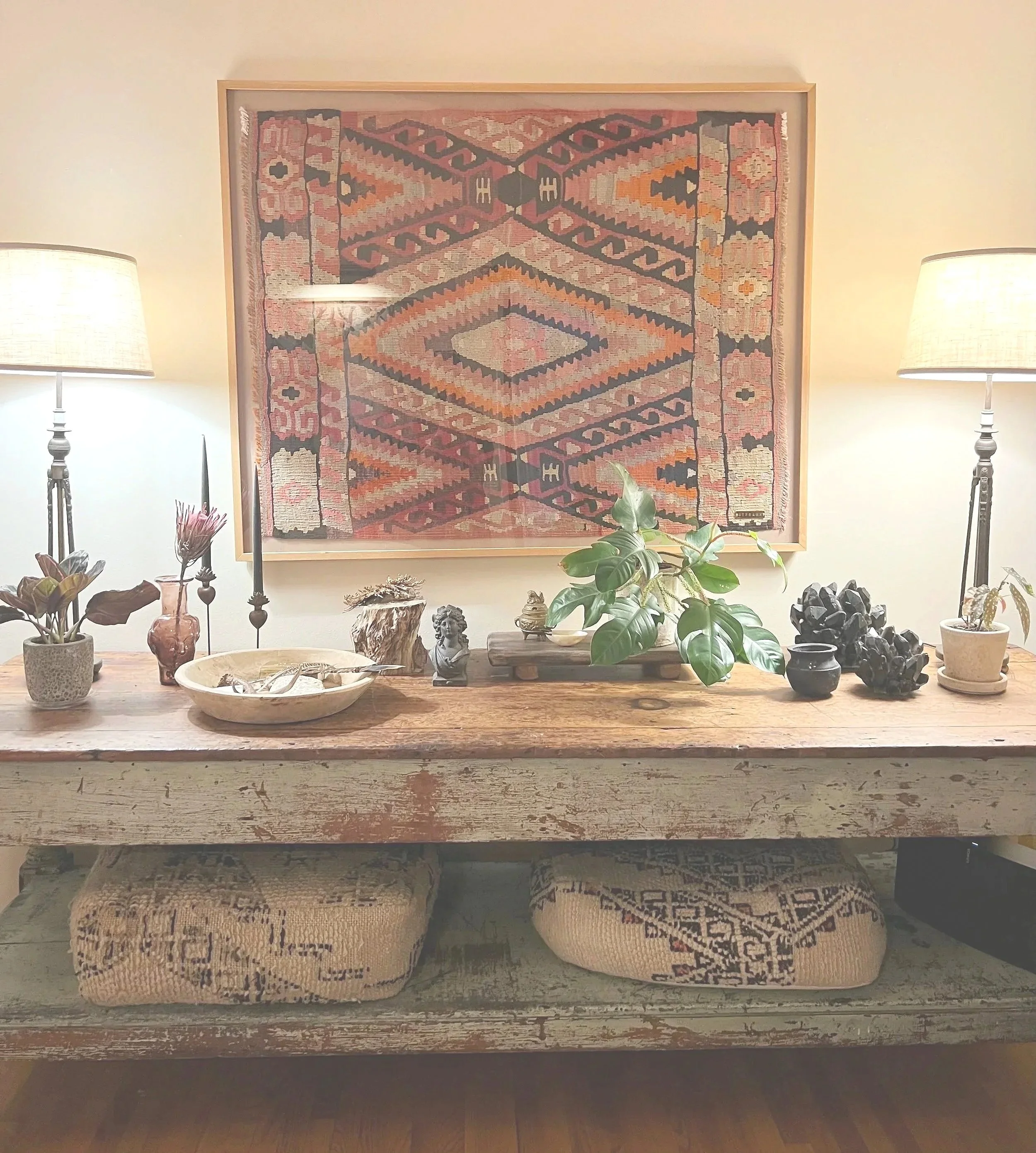 A rustic wooden console table with plants, small sculptures, and decorative objects, topped with a framed colorful geometric-patterned textile, flanked by two table lamps with beige shades.