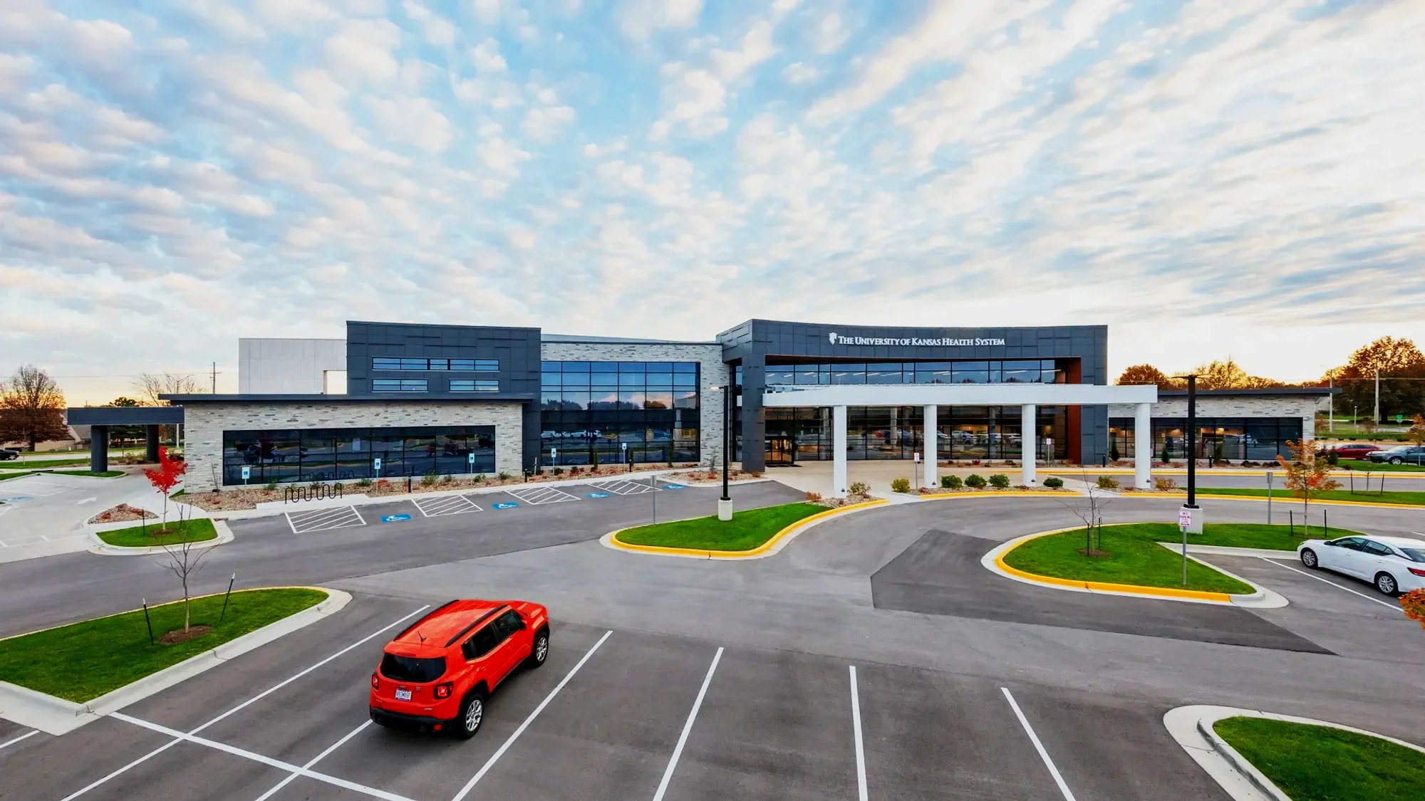 Modern medical facility with parking lot, trees, and a partly cloudy sky. The building has a sign that reads "The University of Kansas Health System."