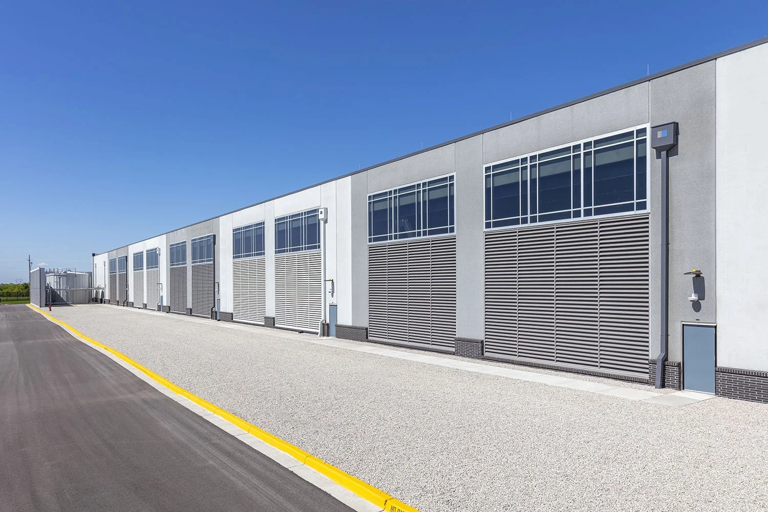 Exterior view of a modern industrial or commercial building with large windows, gray walls, and a yellow curb, under a clear blue sky.