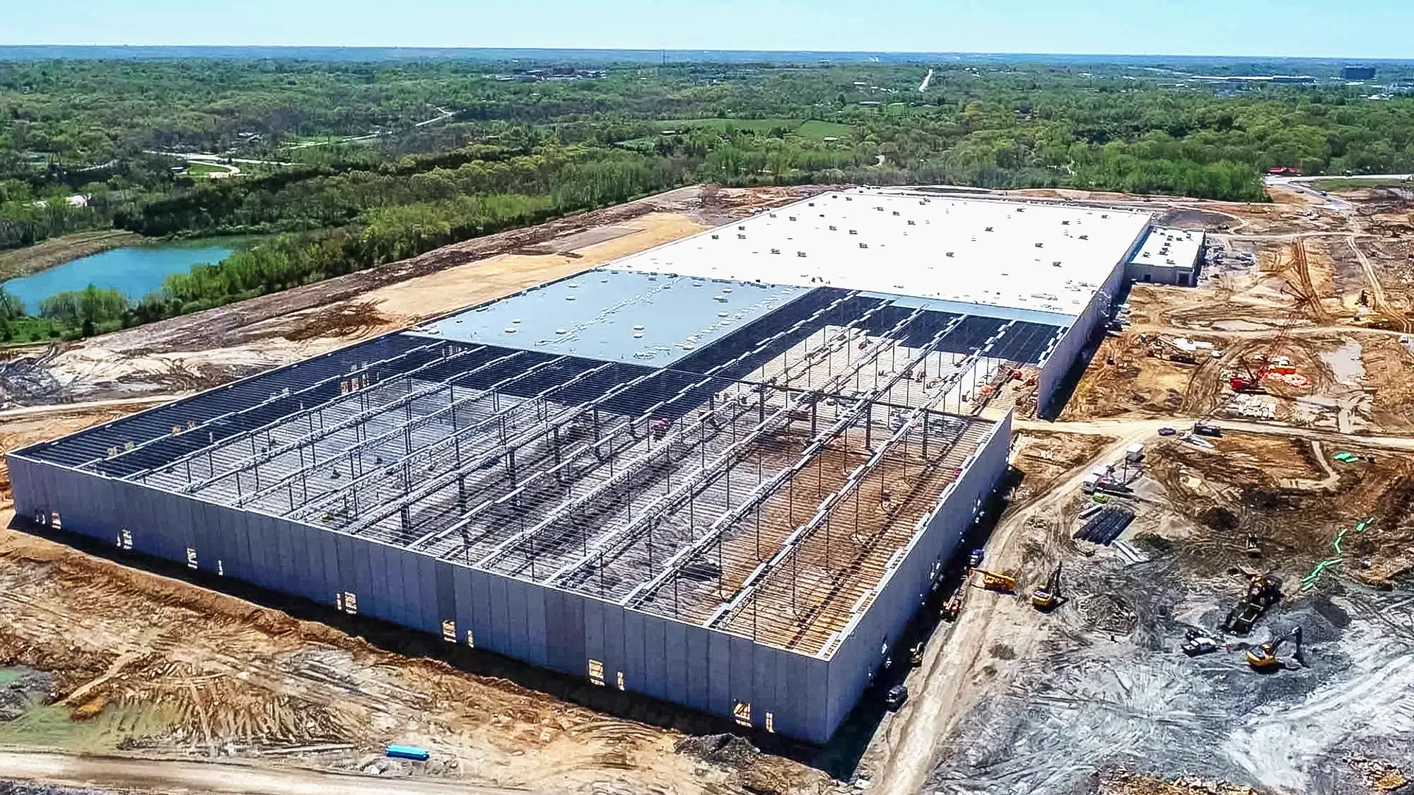 A large building under construction with a steel framework, surrounded by dirt and construction equipment, in a semi-rural area with trees and a pond nearby.