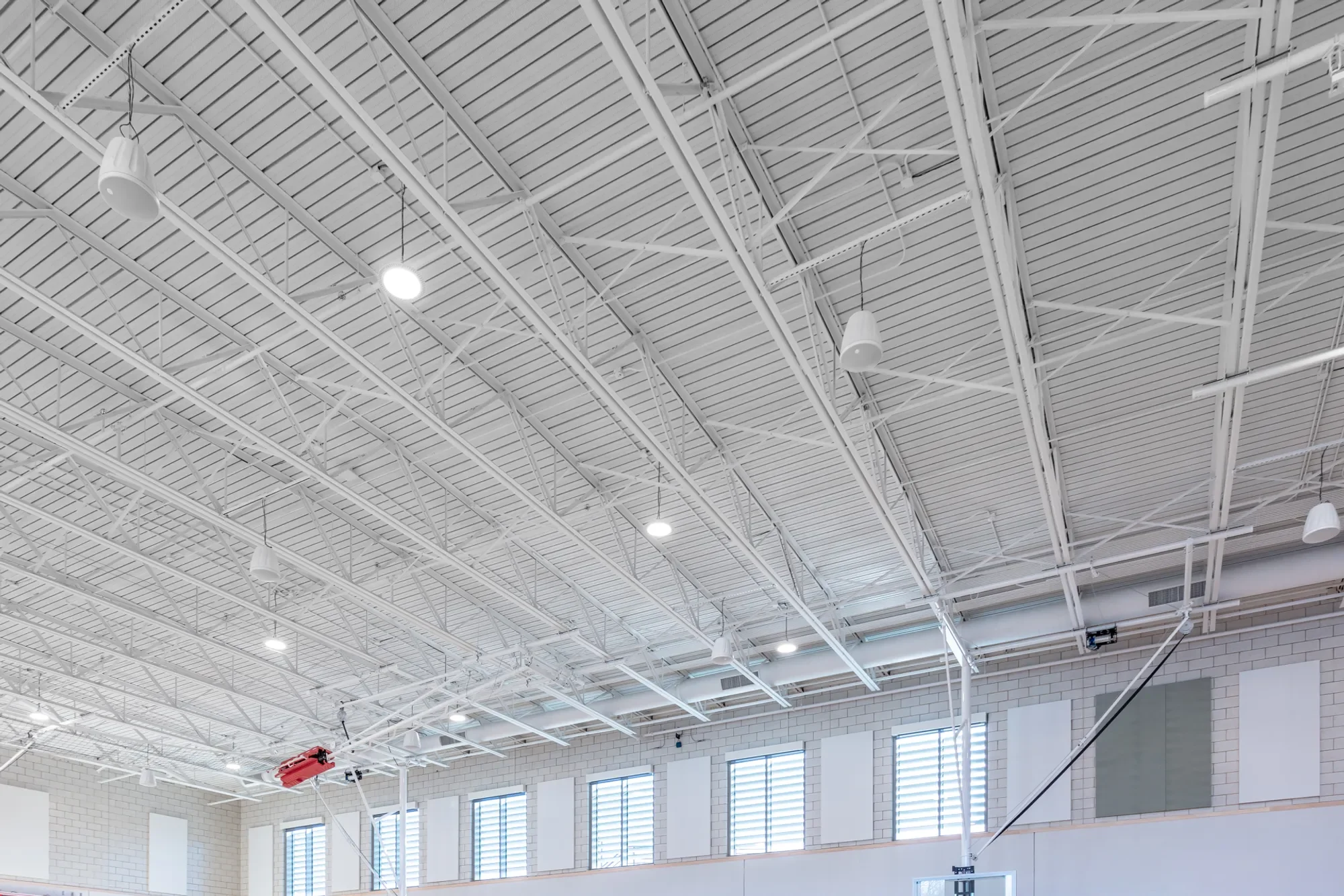 Ceiling of a large indoor space with white structural beams, bright lights, and large windows with blinds.