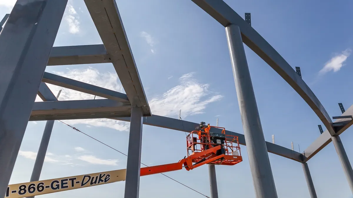 A construction worker on an orange lift working on a steel framework of a building against a blue sky with clouds.