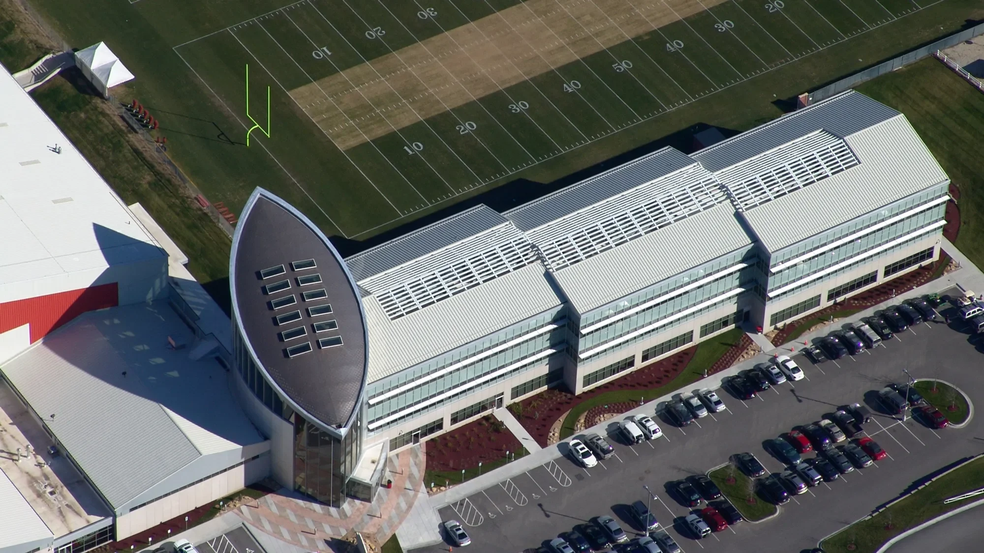 Aerial view of a modern building with a large parking lot, adjacent to a football field with marked yard lines, and a seating area with red seats.
