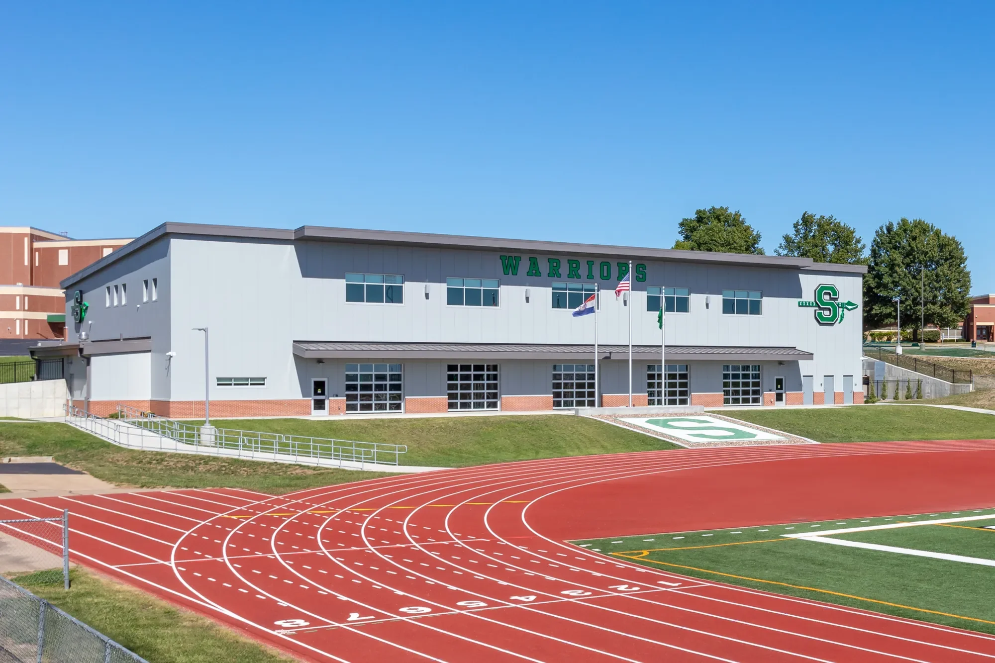 A white athletic building with green 'WARRIORS' lettering and a large 'S' logo on the front, beside a red running track with numbered lanes, along with three flagpoles flying American, Missouri, and green flags, and green trees in the background under a clear blue sky.