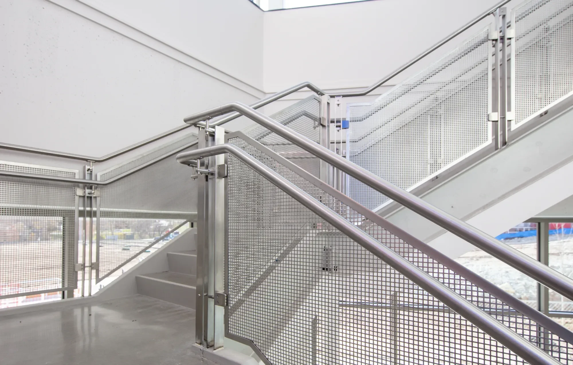 Modern indoor staircase with metal railings and mesh panels, leading to an upper level with large windows and natural light.