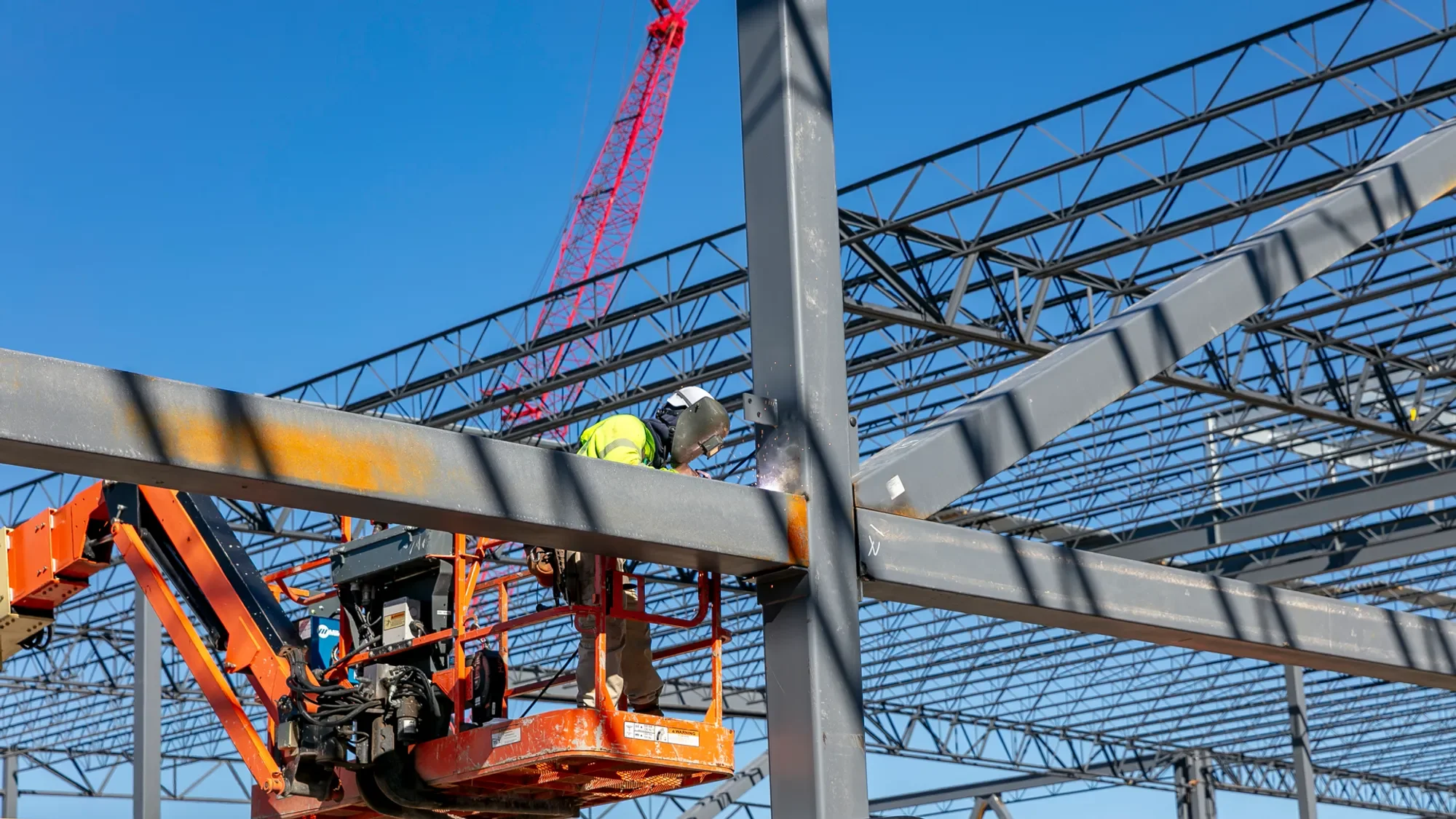 A construction worker wearing a safety helmet and high-visibility vest is welding a metal beam on a construction site. The worker is elevated on an orange lift, with a large crane and steel framework visible in the background under a clear blue sky.