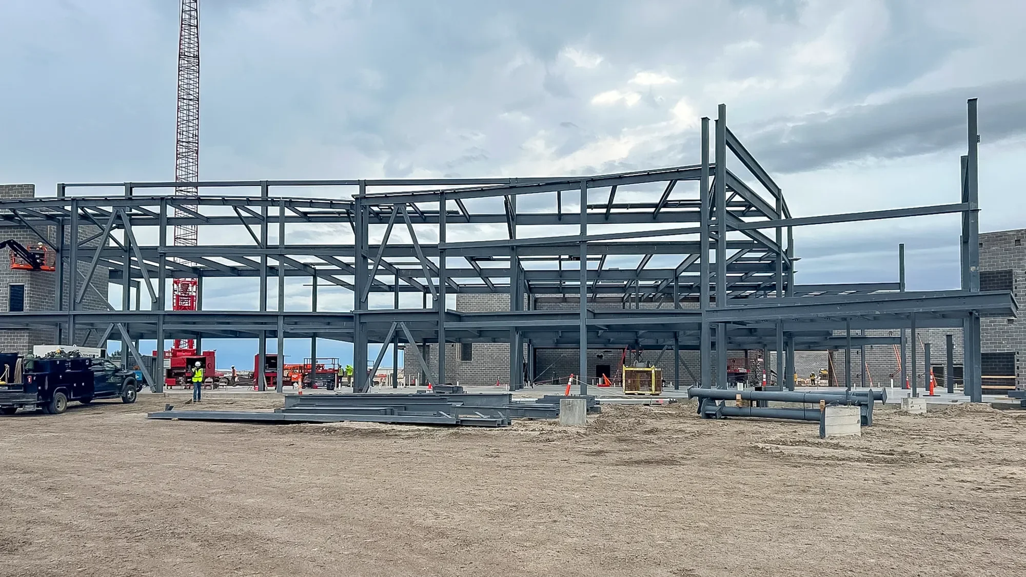 Construction site with steel framework for a building, workers in safety gear, construction equipment, and a cloudy sky.