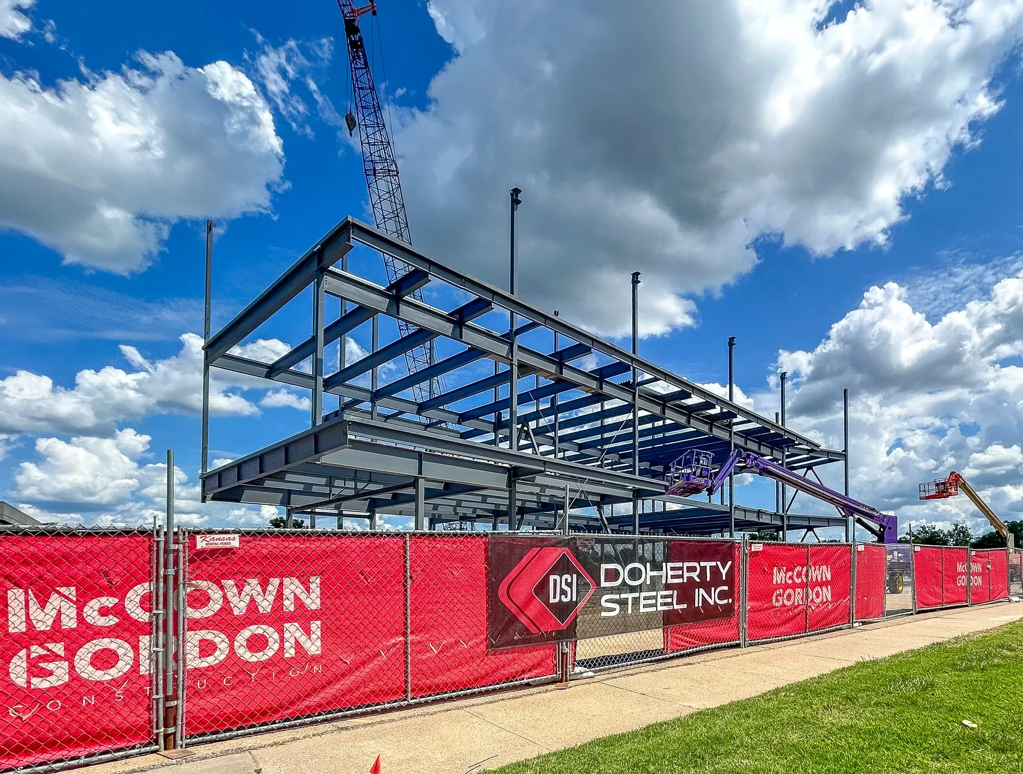 Construction site of a building frame with steel beams and a construction crane against a cloudy blue sky. Red barriers with company logos and a chain-link fence surround the area.