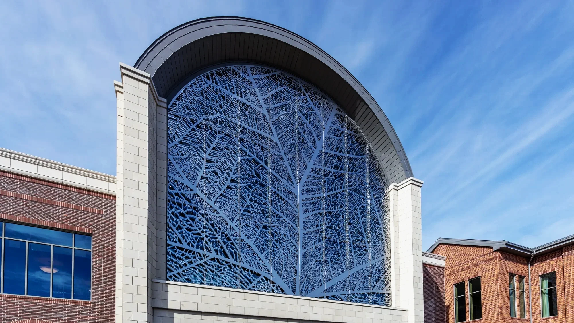 Modern building with a large arched window featuring a blue decorative metal design resembling tree branches, partly made of brick and stone with blue sky in background.