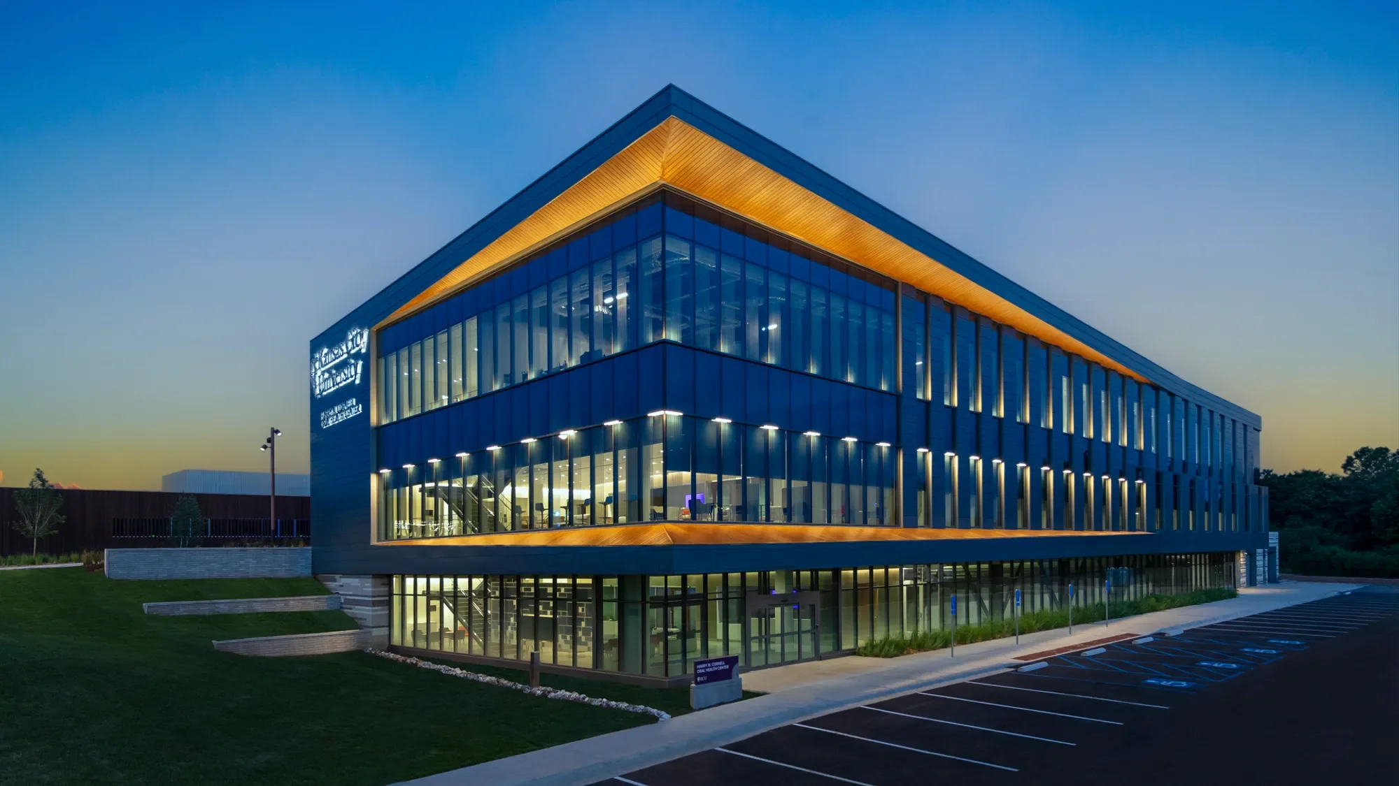Modern multi-story building with glass exterior and yellow accent lighting, parking lot in front, trees in background, during dusk.
