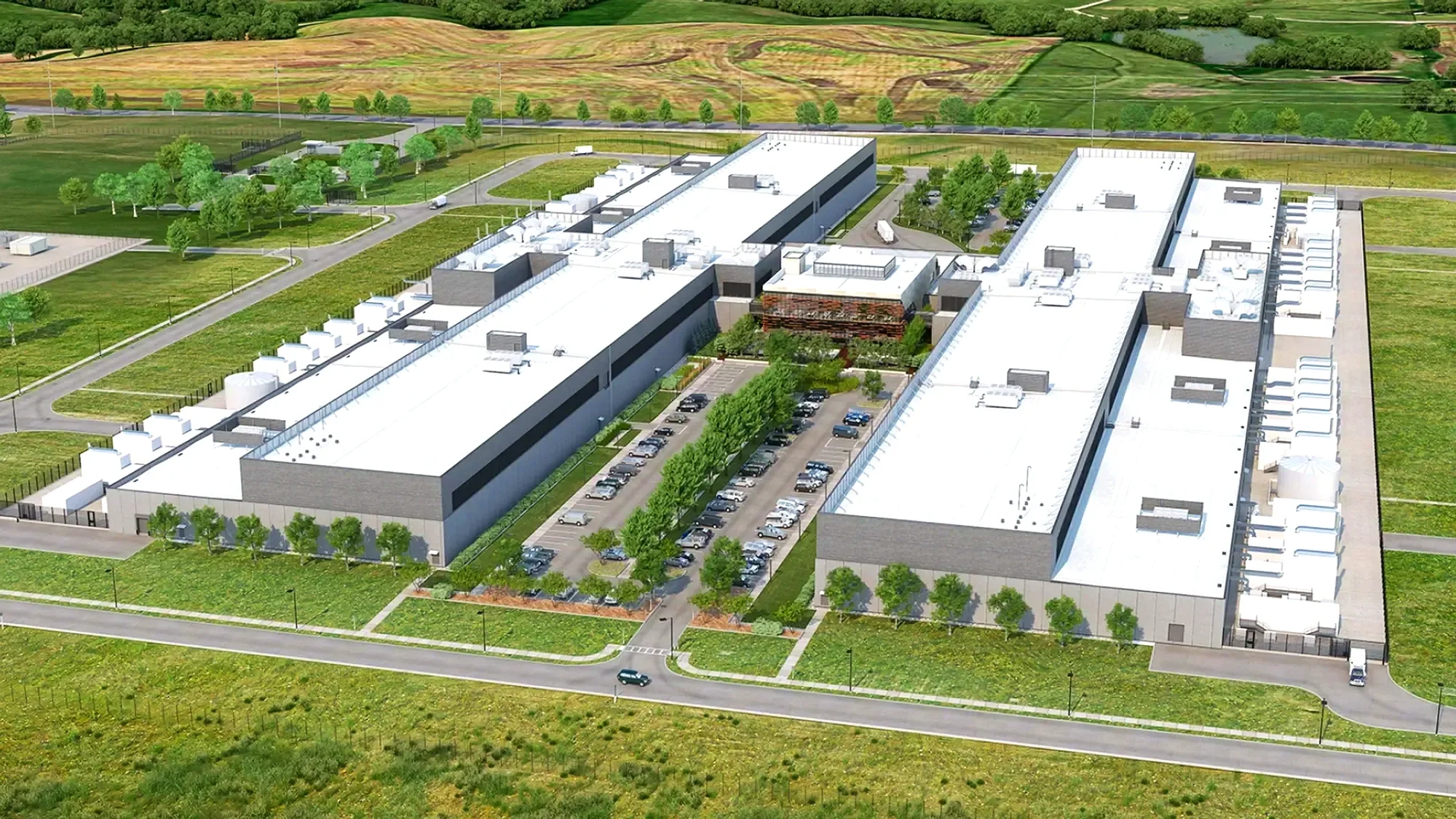 A large modern industrial or commercial building surrounded by parking lots and greenery, with trees, grass, and a distant landscape of rolling hills and fields.