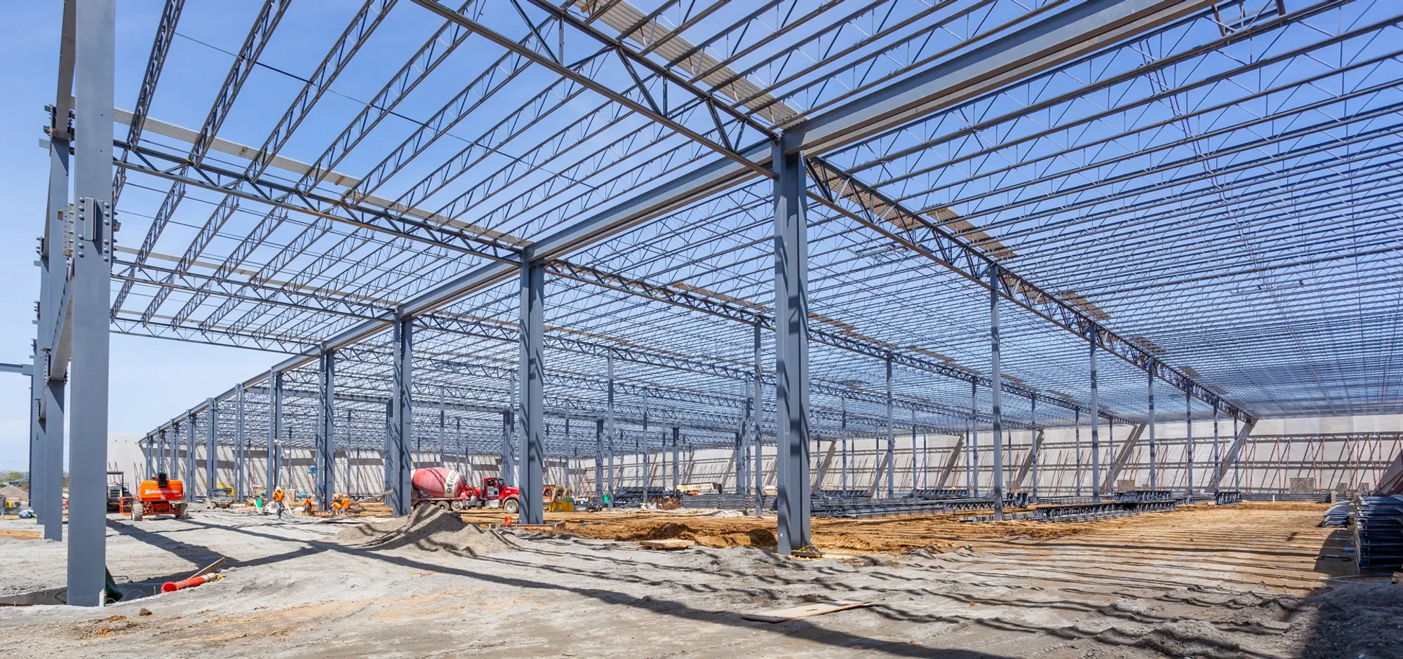 Construction site of a large industrial building with steel framework and a partially completed roof, workers and construction equipment on site.