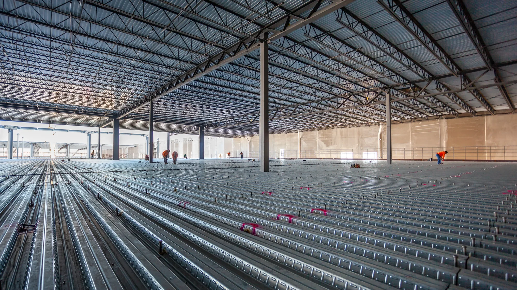 Interior view of a large industrial building under construction with steel beams and framework, with workers installed on the floor during the construction process.