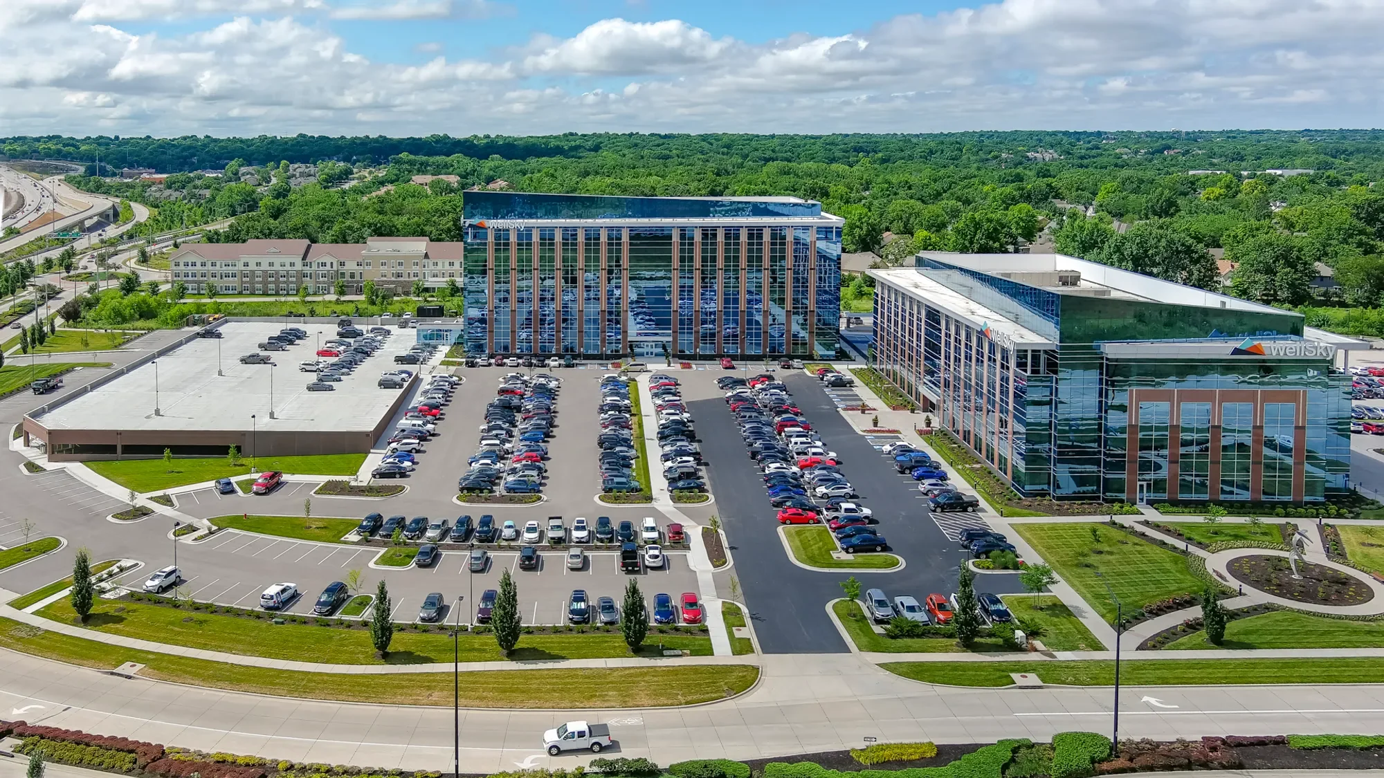 An office building with reflective glass windows and a large parking lot in front. The area is surrounded by greenery and trees, with a highway in the background under a partly cloudy sky.