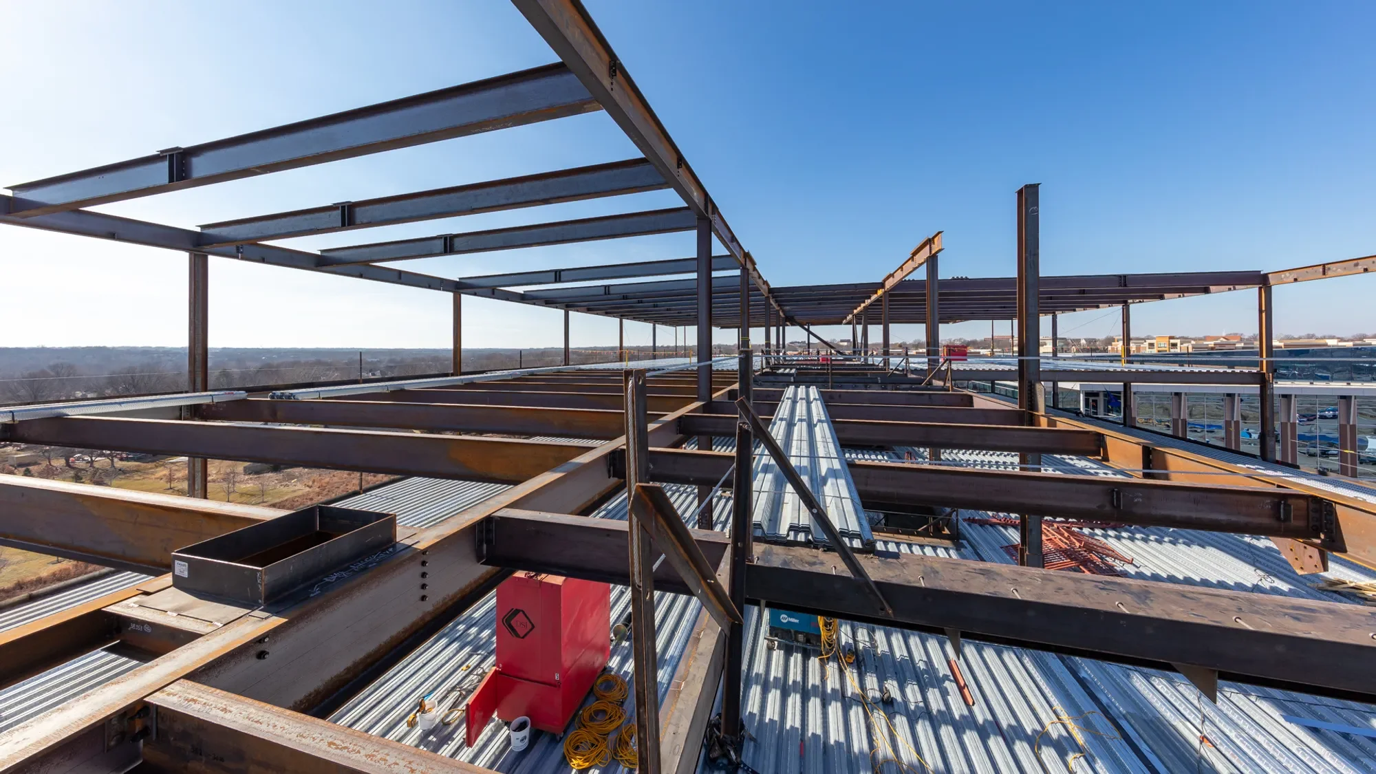 Construction site on the roof of a building with a steel frame structure and metal roofing panels, with a view of the surrounding area under a clear blue sky.