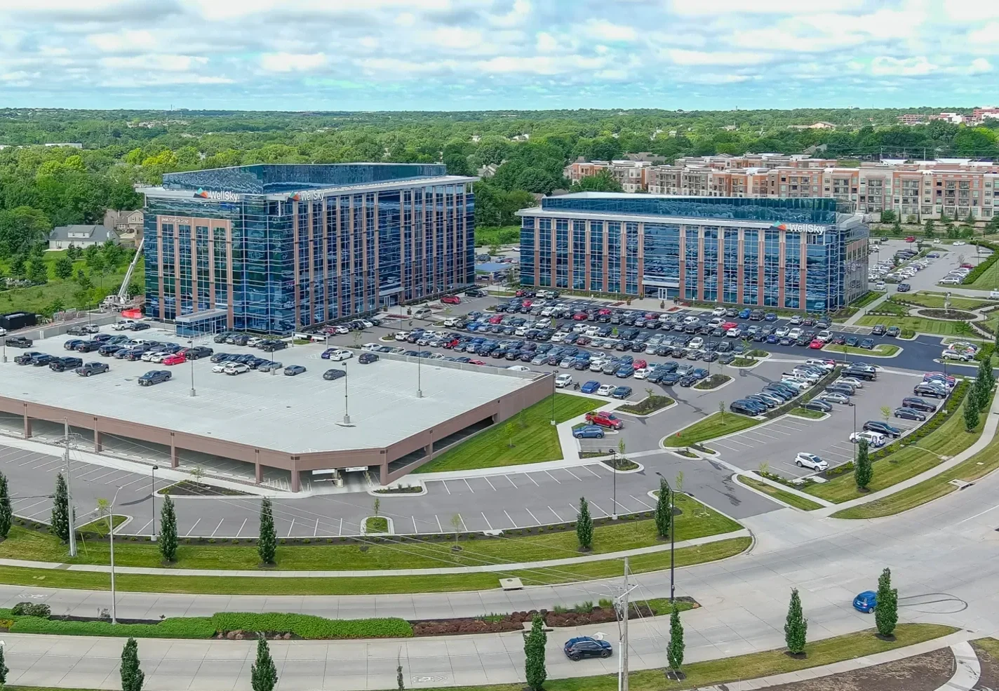 Aerial view of two modern glass office buildings with parking lots, surrounding roads, greenery, and a residential area in the background.