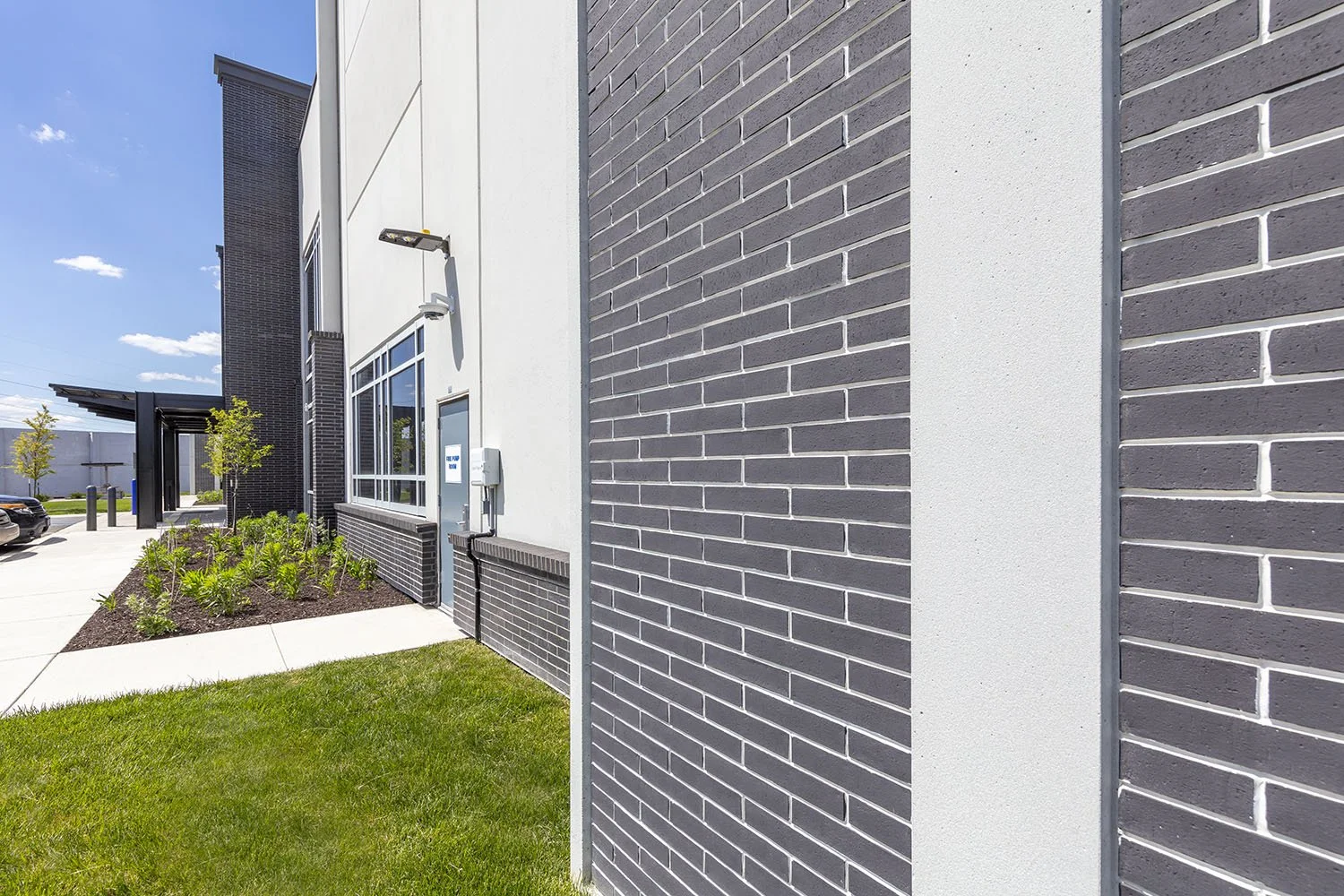 Exterior view of a modern commercial building with dark and white brick walls, large windows, a small landscaped area with plants, a sidewalk, and a parking lot under a clear blue sky.