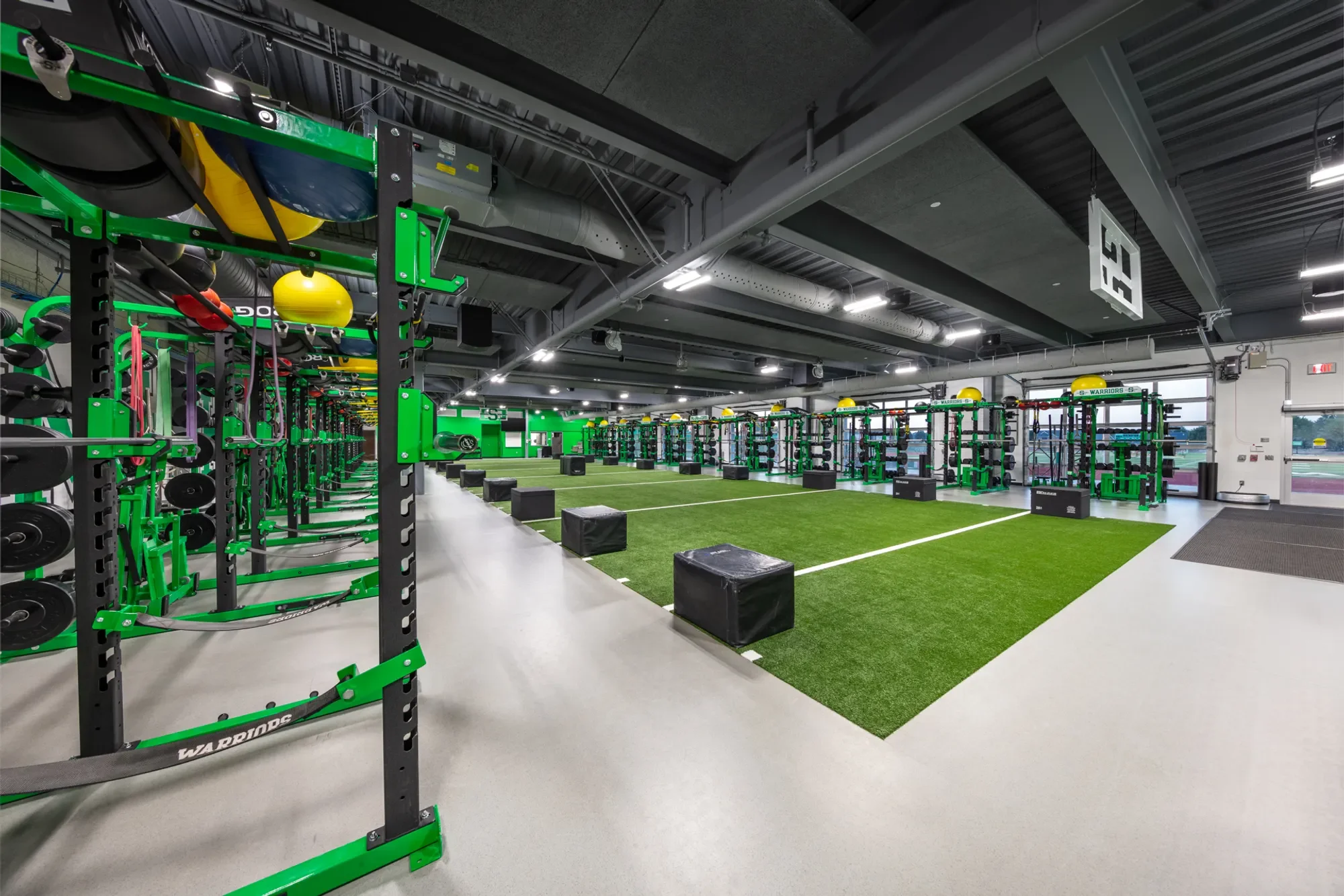 Inside a modern gym featuring black and green workout equipment, a turf area with black plyometric boxes, and a ceiling with industrial style ducts and lighting.