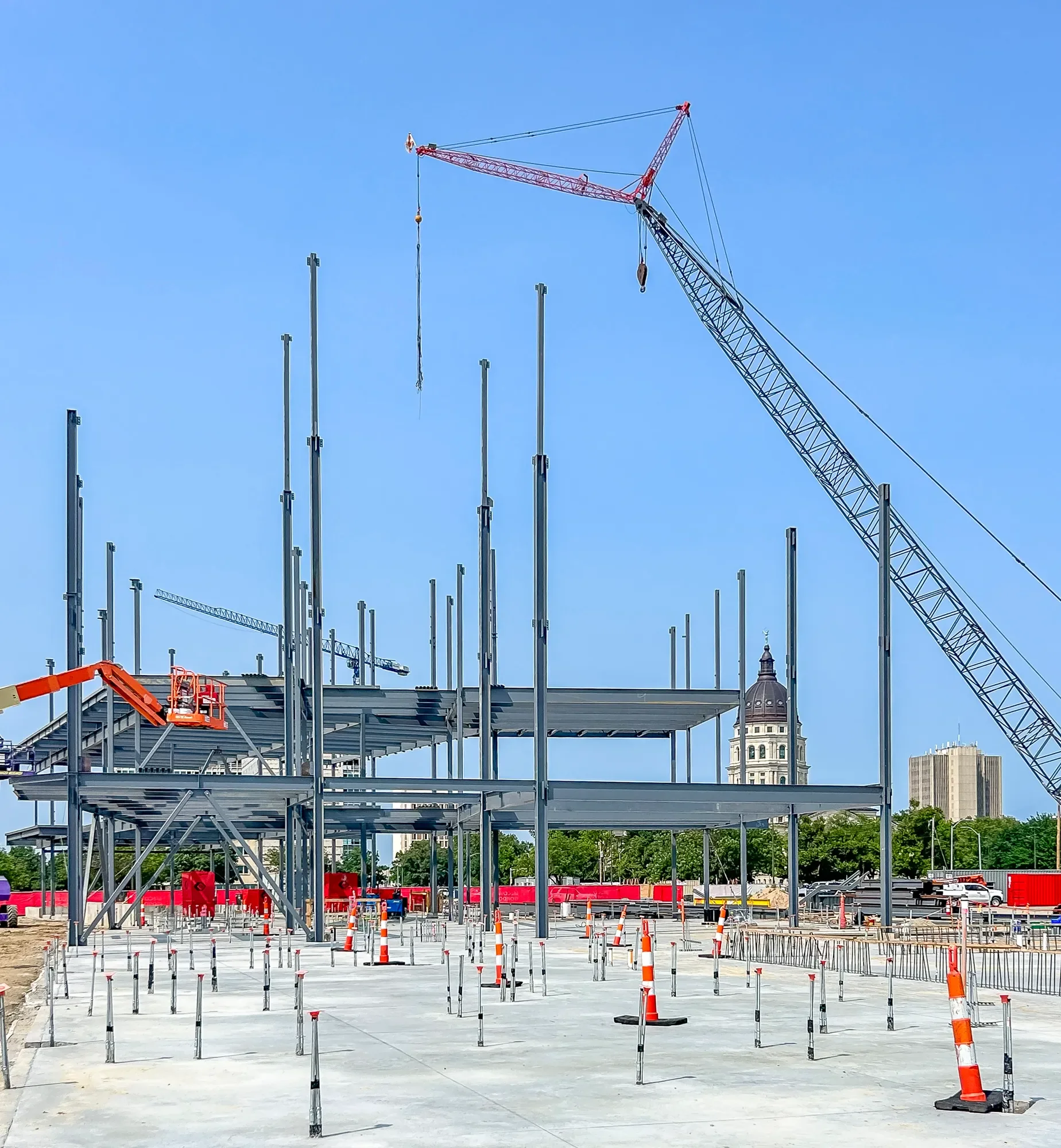 Construction site with steel framework being assembled, orange and white safety cones, a crane lifting materials, and a city skyline with historic buildings and modern skyscrapers in the background.