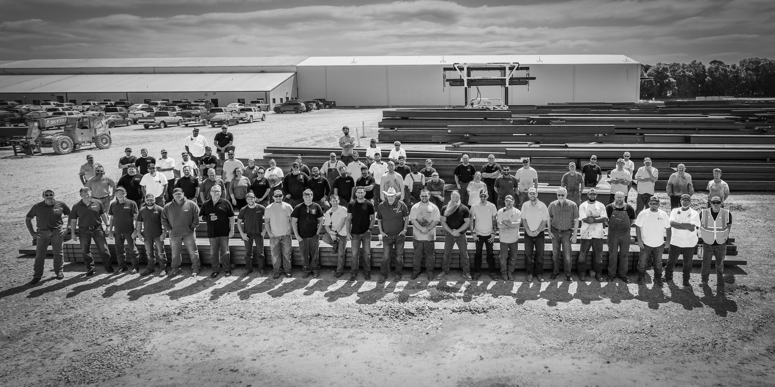 A large group of people standing outdoors in front of stacks of metal beams, with a warehouse and parked cars in the background on a clear day.