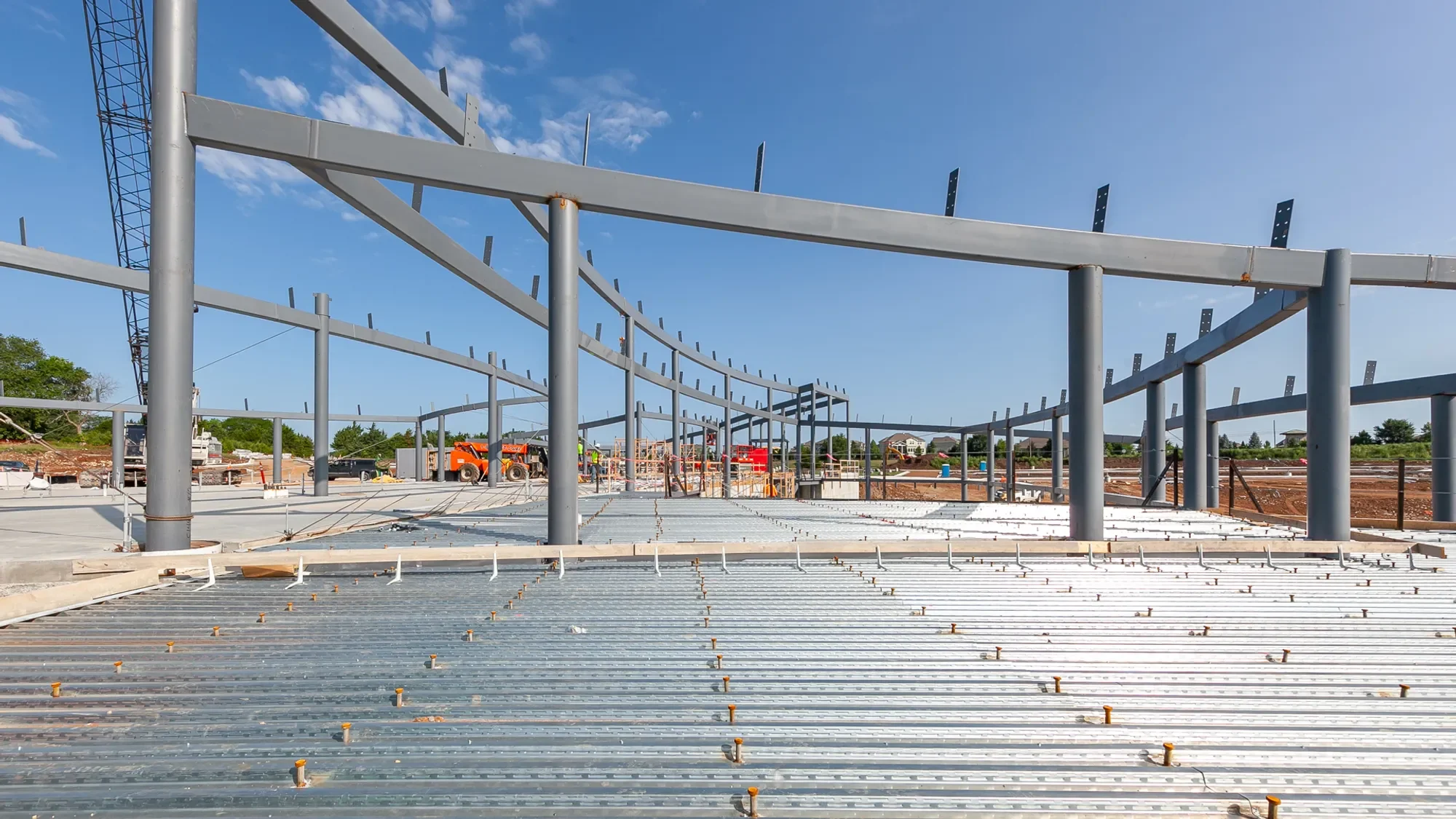 Steel framework of a building under construction with workers and construction equipment in the background under a blue sky.