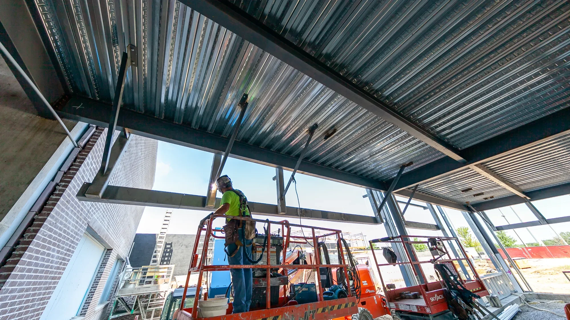 Construction workers on a scissor lift installing a metal roof over a commercial building, with building material and equipment visible on site.