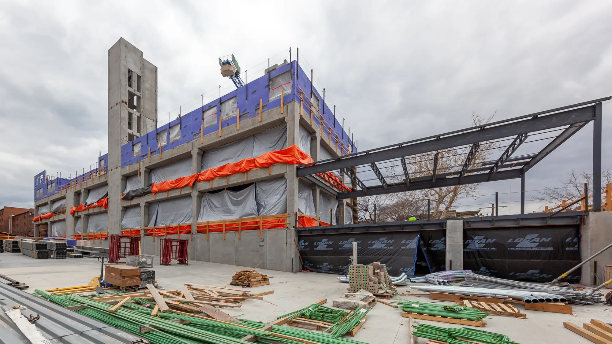 The image shows a building under construction with concrete structure and scaffolding, with construction materials and tools scattered around the site on the ground.
