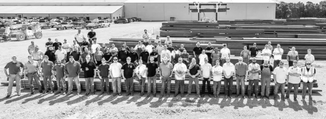 A group of people standing outdoors in front of a large industrial building and parked cars