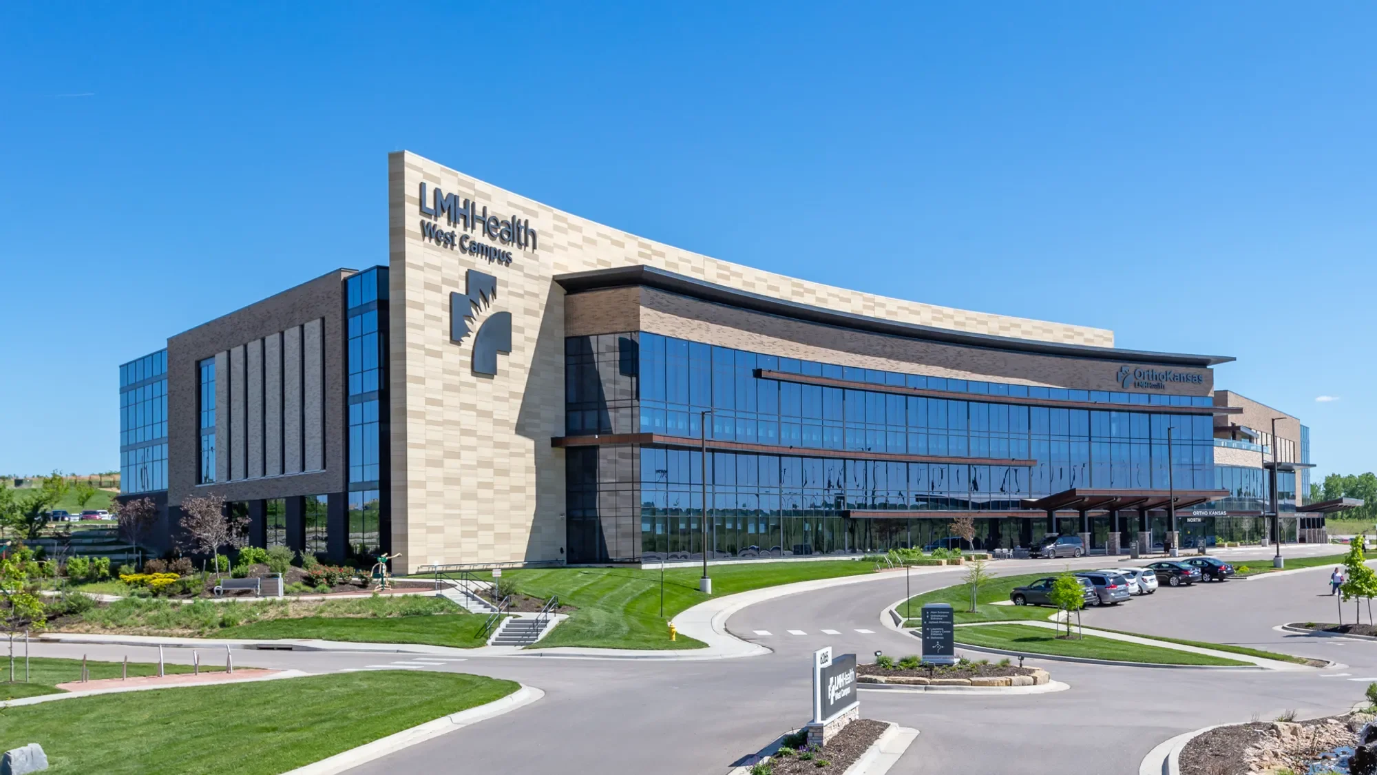 Modern hospital building with blue glass windows, a sign reading 'LMH Health West Campus,' and a parking lot with several cars surrounding it, under a clear blue sky.
