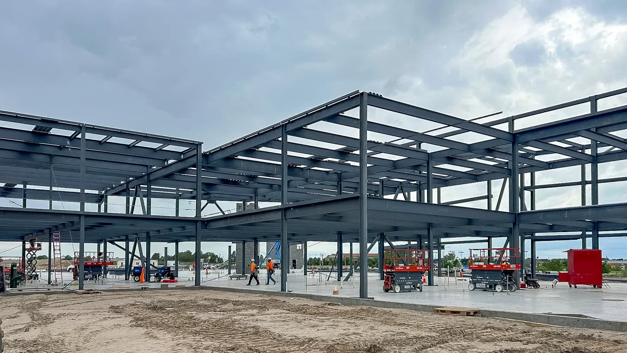 Steel framework of a building under construction at a commercial site with construction workers and equipment, cloudy sky in the background.