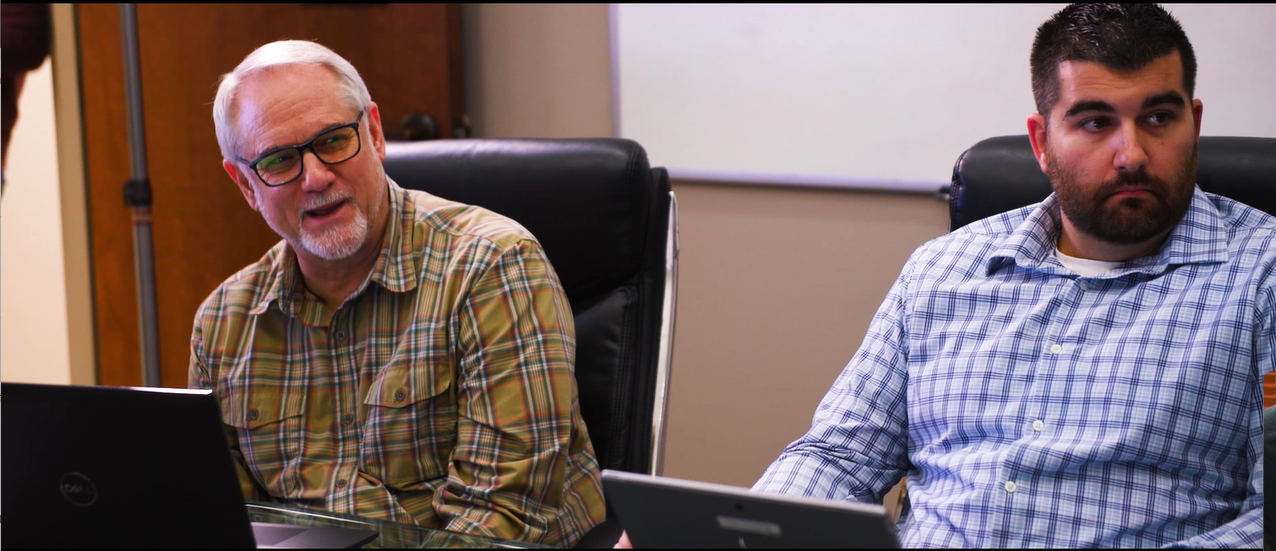 Two men seated at a conference table in a meeting room, one with gray hair and glasses talking, the other with a beard listening, in front of laptops.