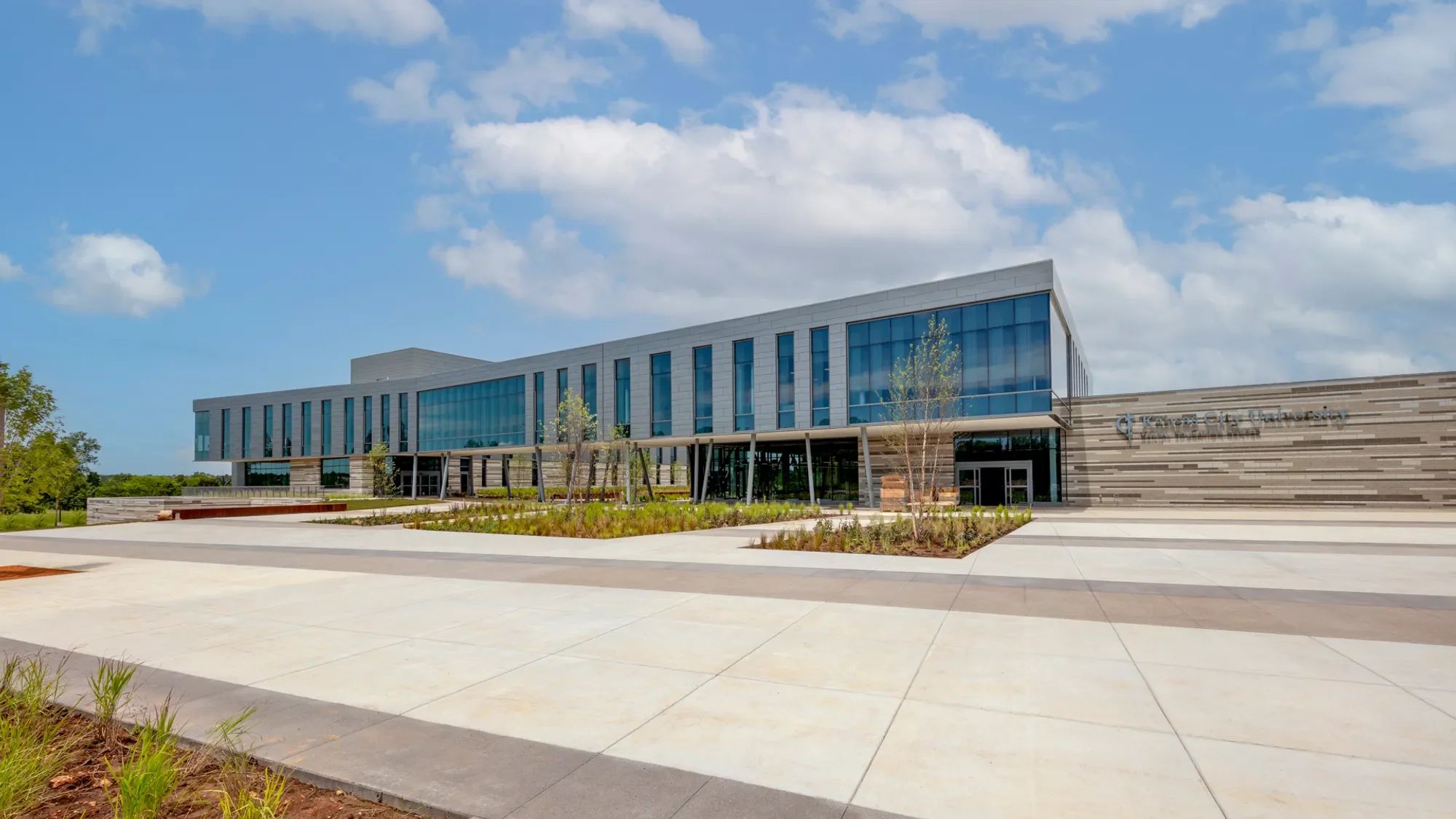 Exterior view of Kansas City University building with modern architecture, large glass windows, pathway, and landscaped area with small trees and plants under partly cloudy sky.