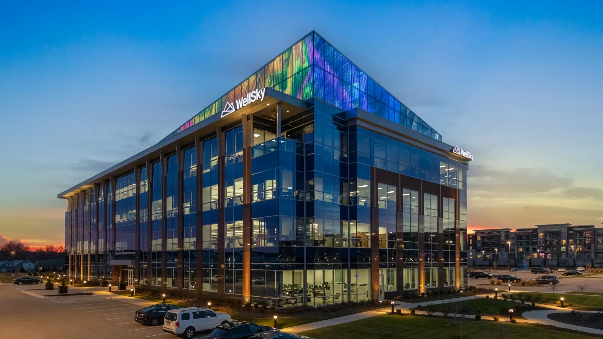 Modern glass office building with illuminated interior, featuring the Wellsky logo at sunset with a parking lot in the foreground.