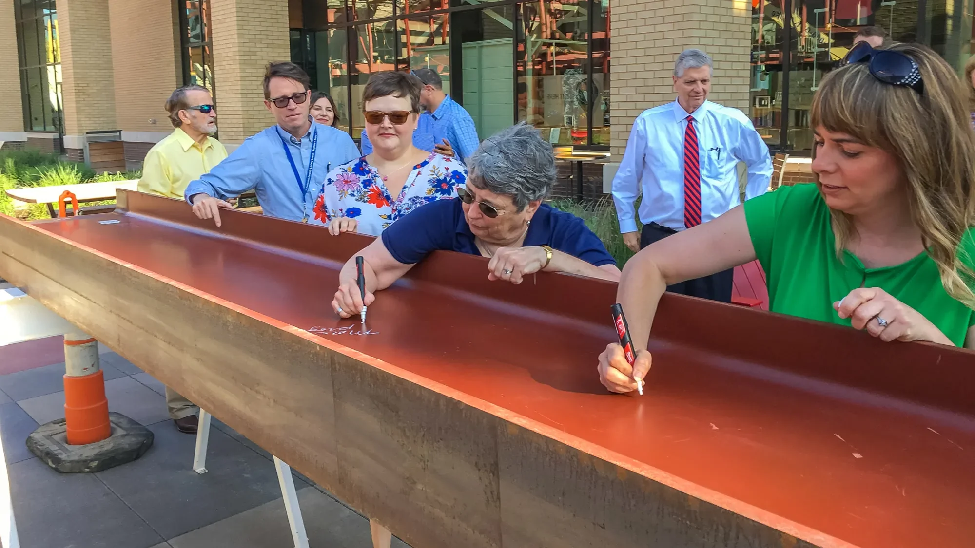 People gathered around a sidewalk game of shuffleboard, with some players writing on the board and others observing, outside in an urban area.