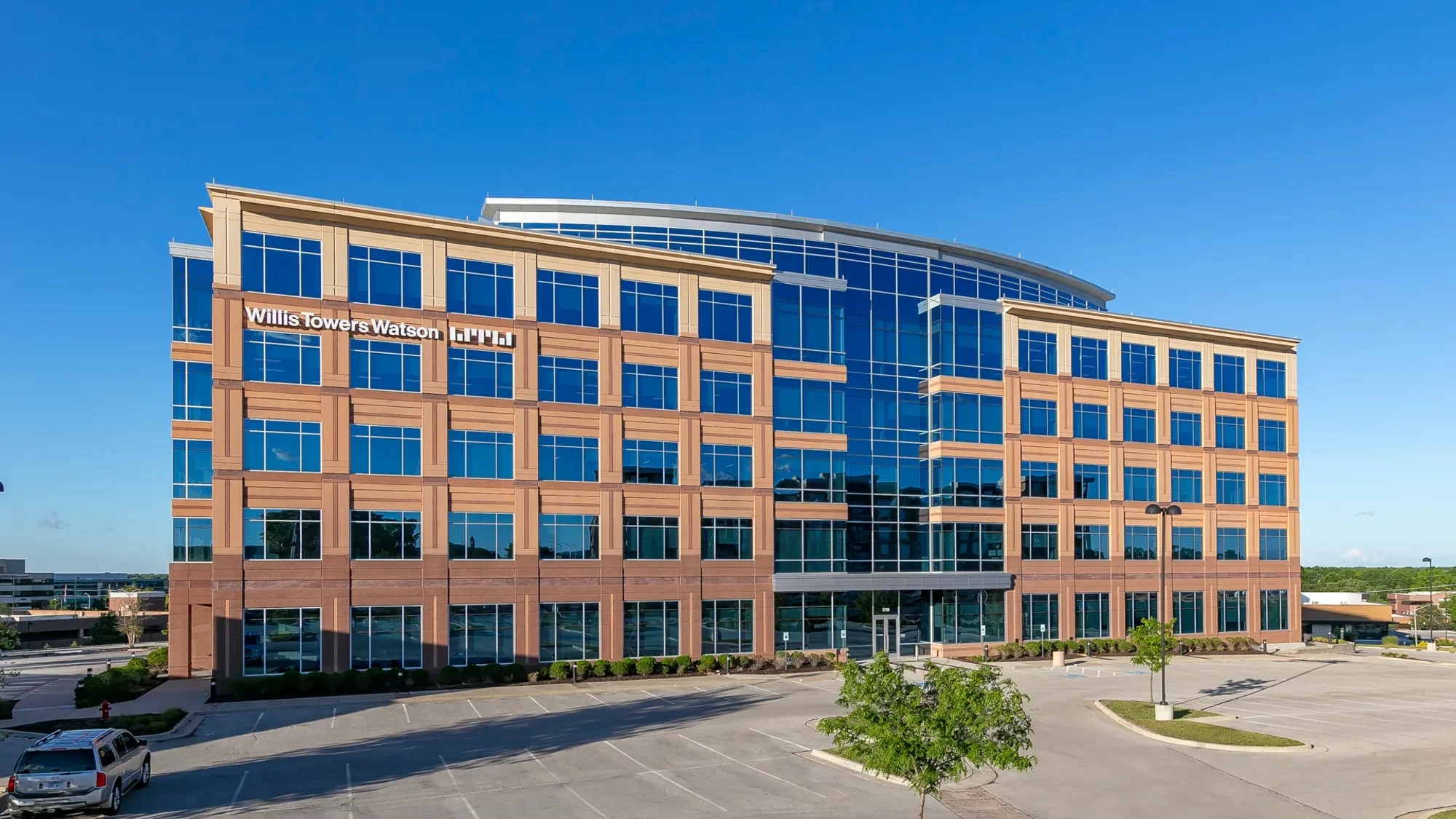 Modern multi-story office building with large blue glass windows, labeled as Willis Towers Watson, with an empty parking lot and few trees in the foreground, under a clear blue sky.