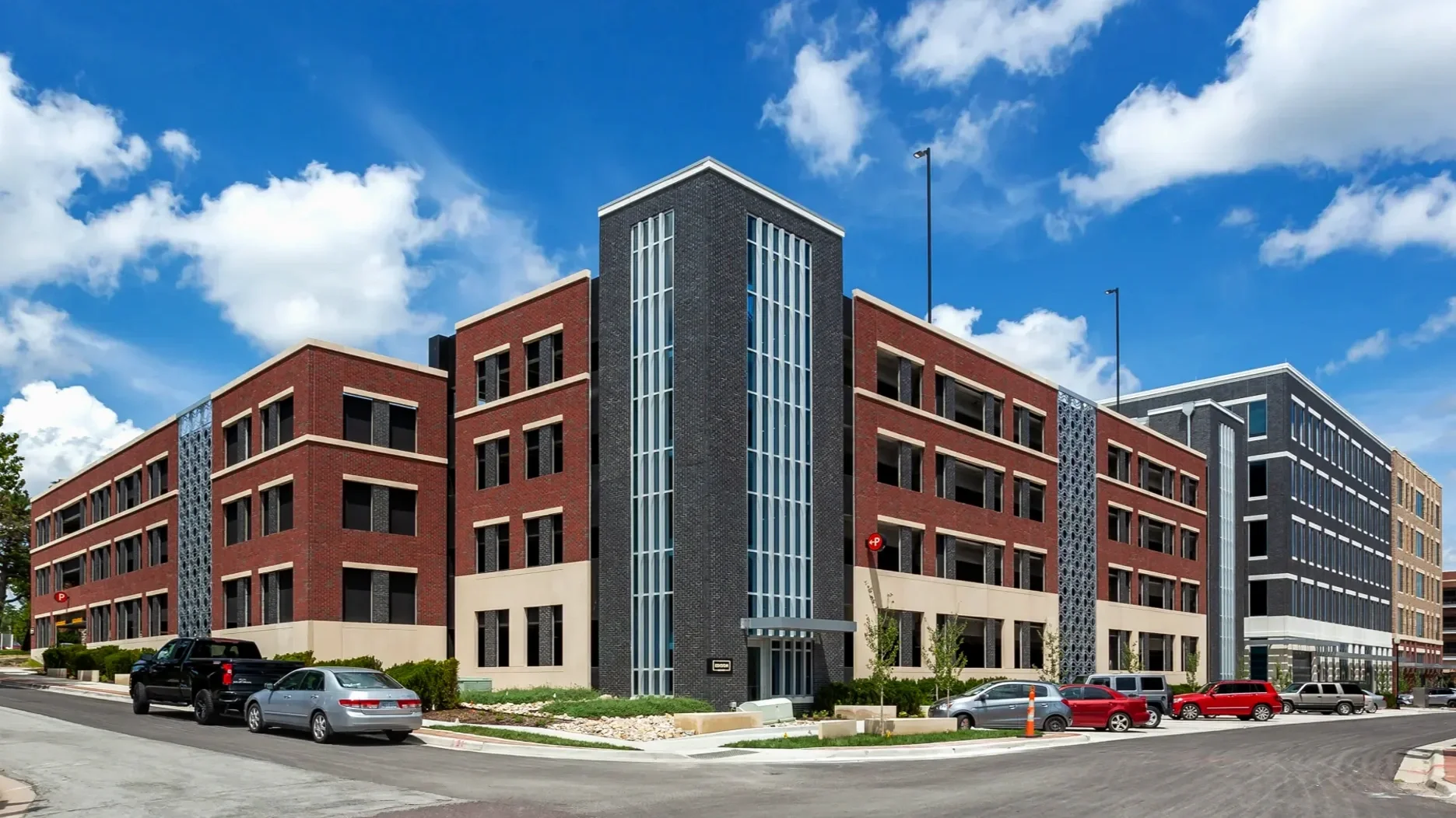 A modern multi-story building with a red and black brick facade, large vertical windows, and a central glass tower, under a partly cloudy blue sky.
