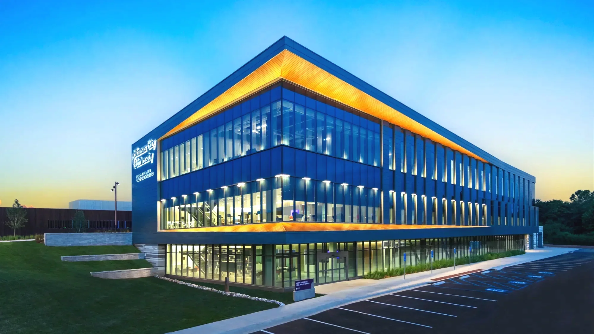Modern multi-story office building with glass exterior and orange accent lighting on the roof, illuminated at dusk, with parking lot in the foreground and a green lawn surrounding the building.