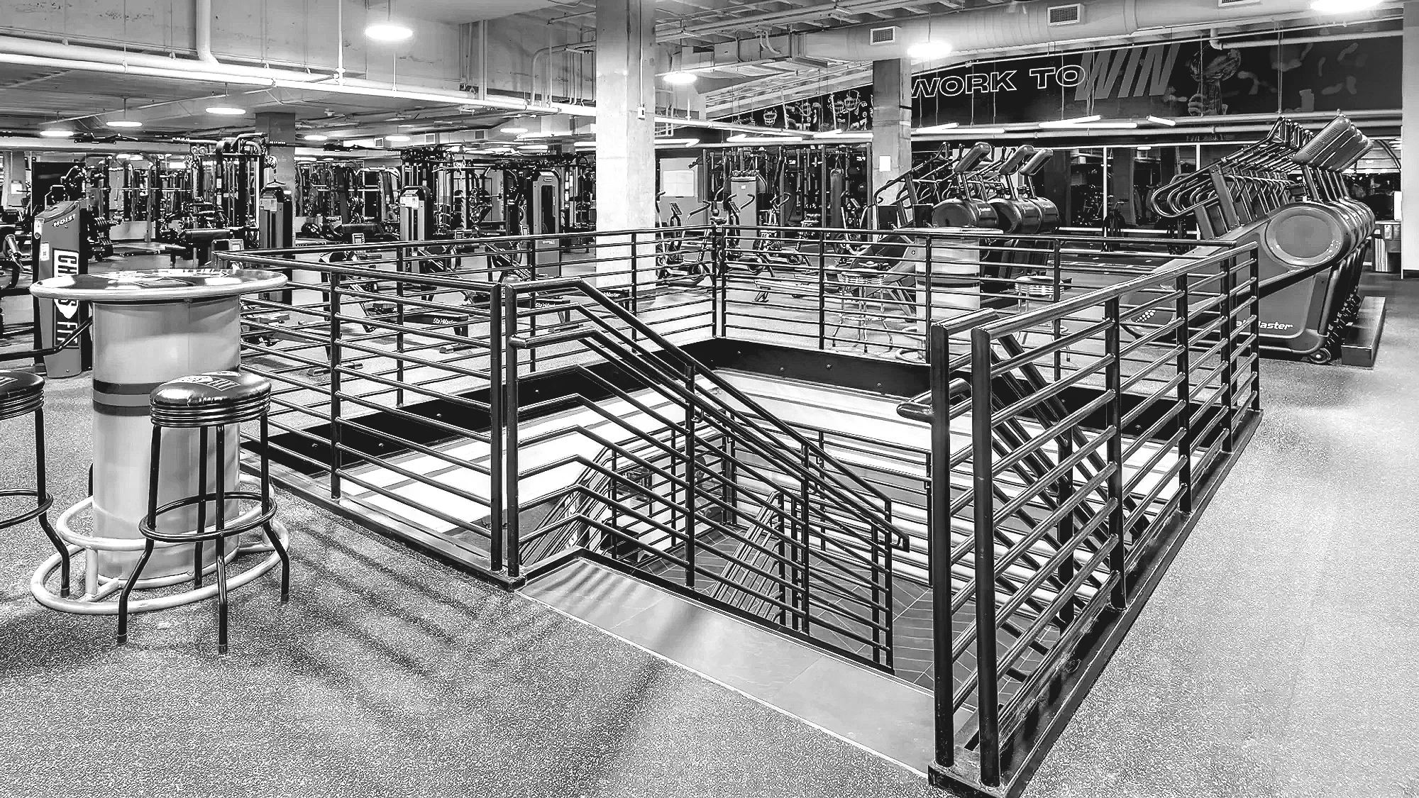 Empty fitness gym with weightlifting equipment, stationary bikes, and a staircase with black railing in black and white.