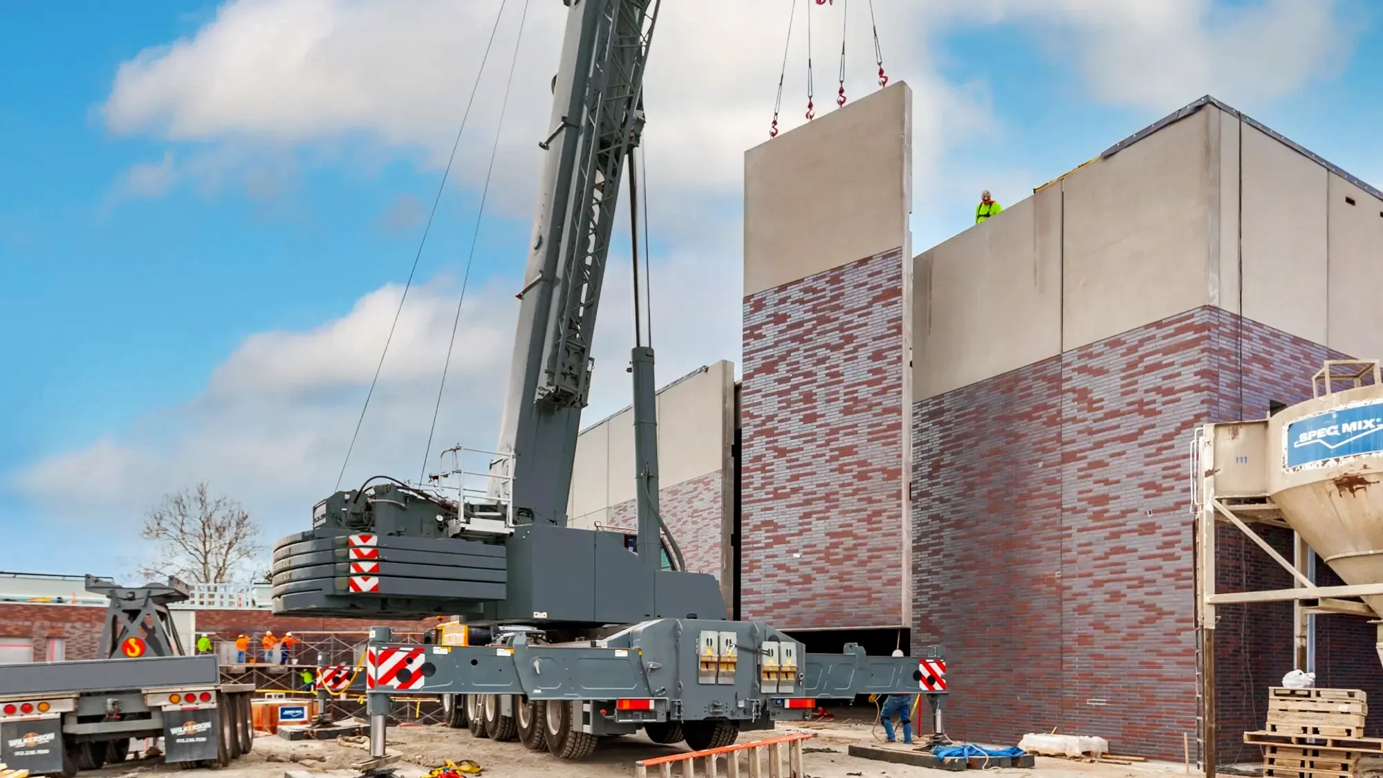Construction site with a large crane lifting a wall section of a building with brick accents, workers in safety gear, and construction equipment in the foreground.
