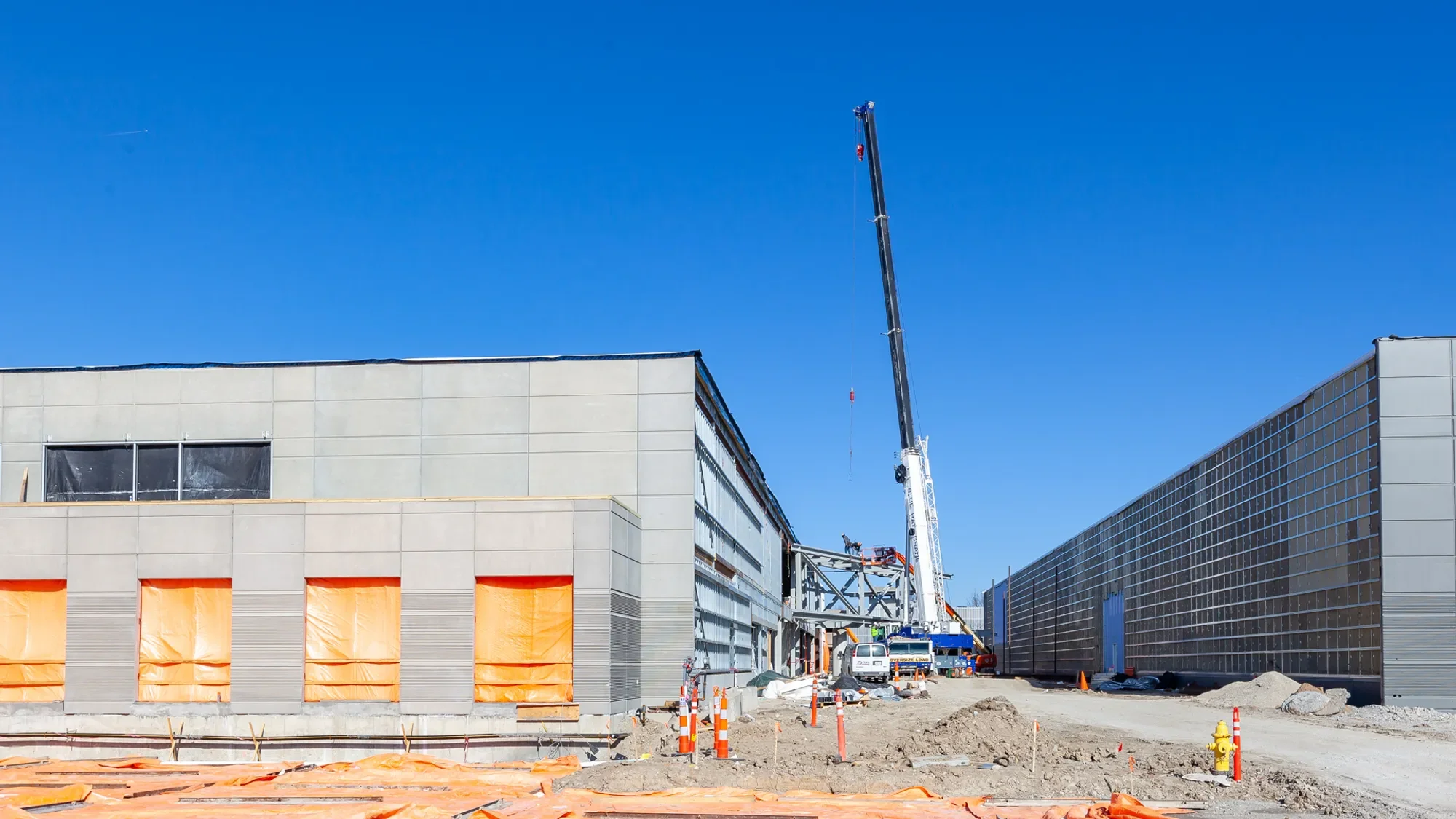 Construction site with a crane and modern buildings, some windows are covered with orange protective films, ground is unfinished with construction materials and orange safety barriers.