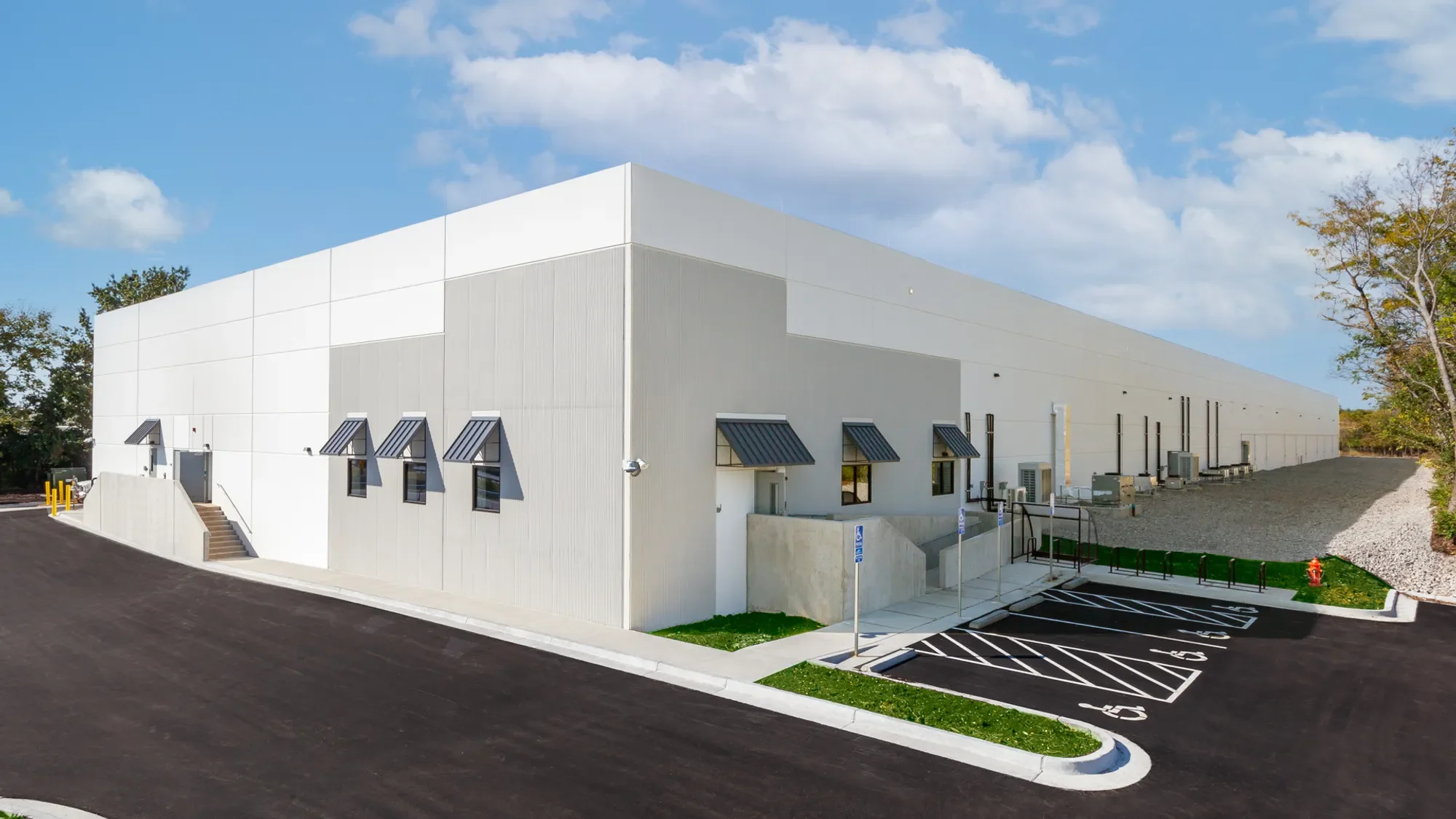A large white commercial or industrial building with a flat roof, small windows with metal awnings, and multiple air conditioning units outside. There is a parking lot with designated parking spaces, including accessible parking, and a few trees in the background under a partly cloudy blue sky.