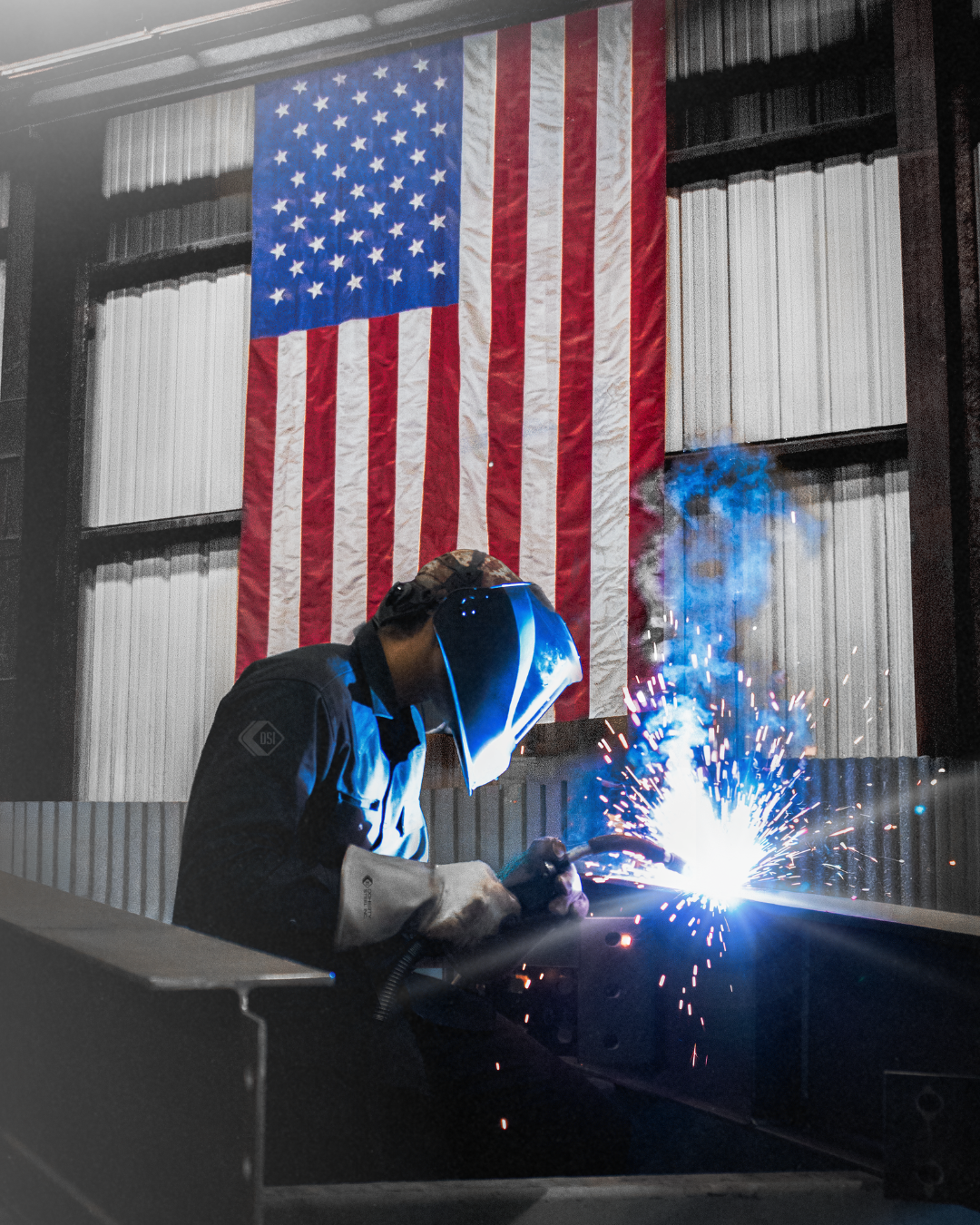 A welder wearing protective gear and a welding helmet welding metal with bright sparks, in front of an American flag hanging in an industrial setting.