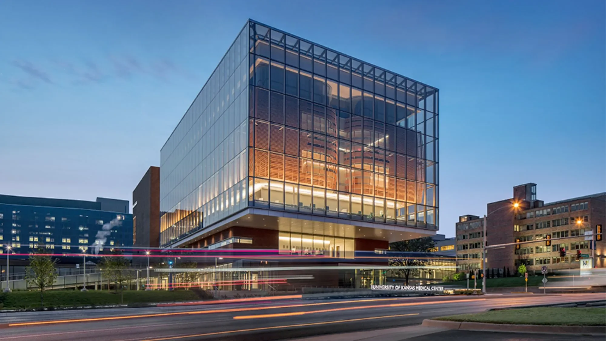A modern glass building, the University of Kansas Medical Center, illuminated at dusk with city and traffic lights in the background.