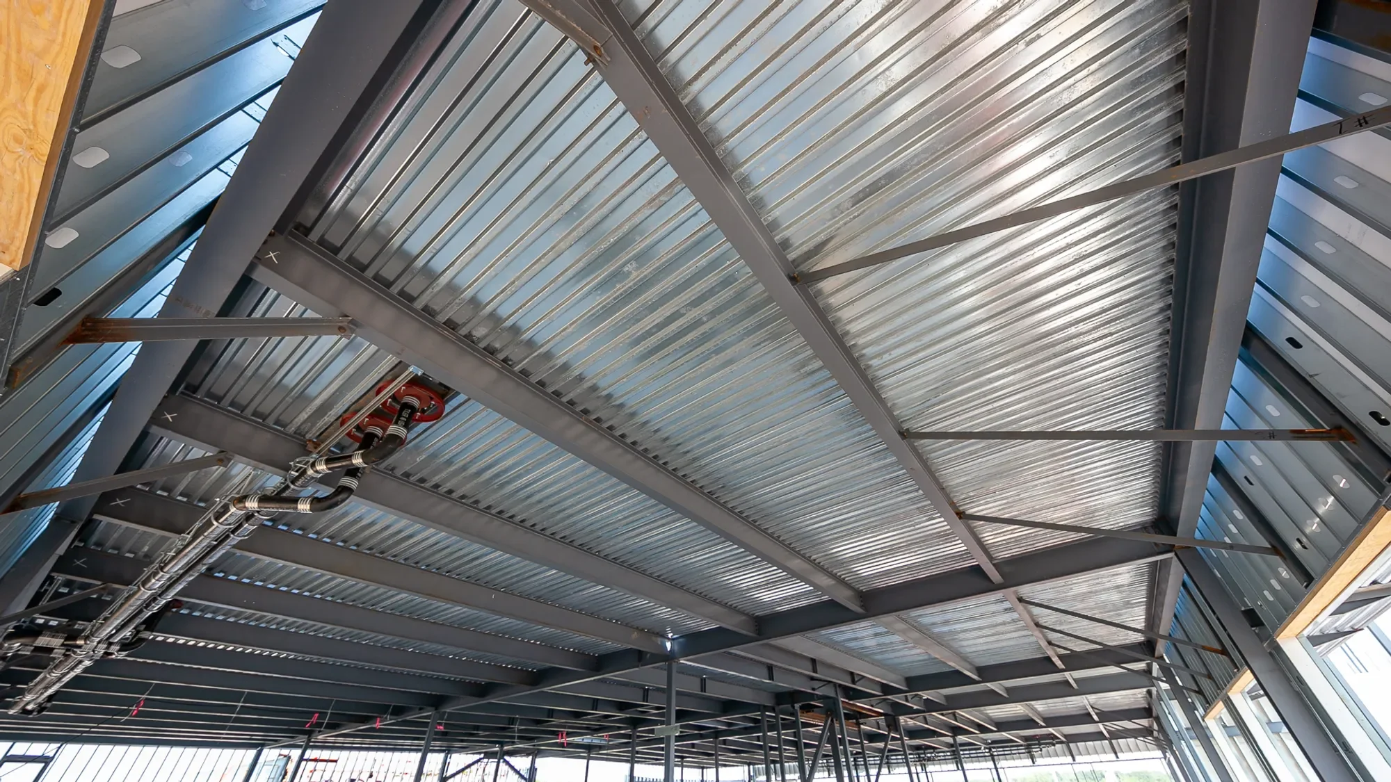 Construction site ceiling with metal beams, corrugated metal panels, and exposed piping.