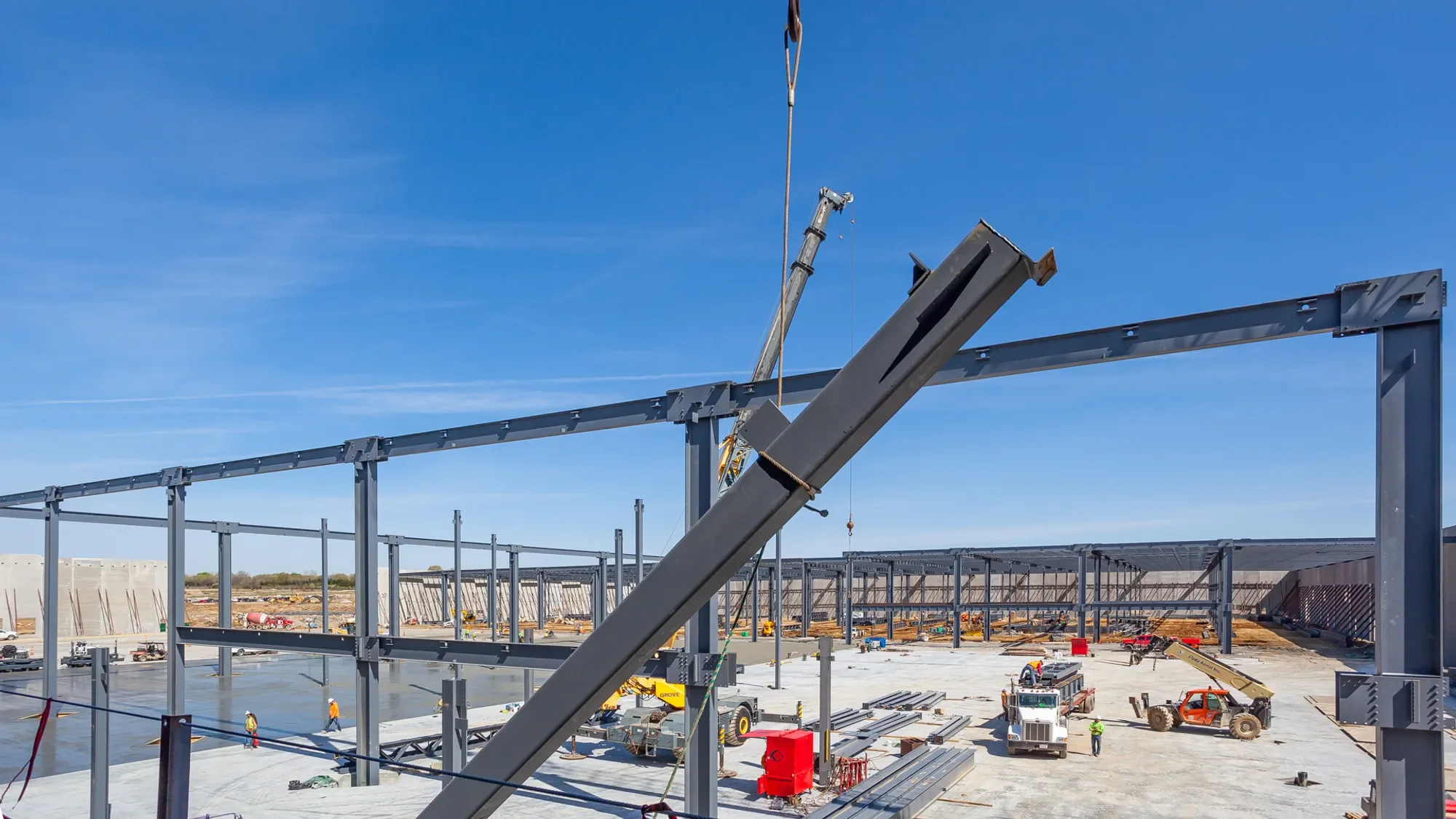 Construction site with steel framework and workers, crane lifting steel beam, machinery, and equipment under a clear blue sky.