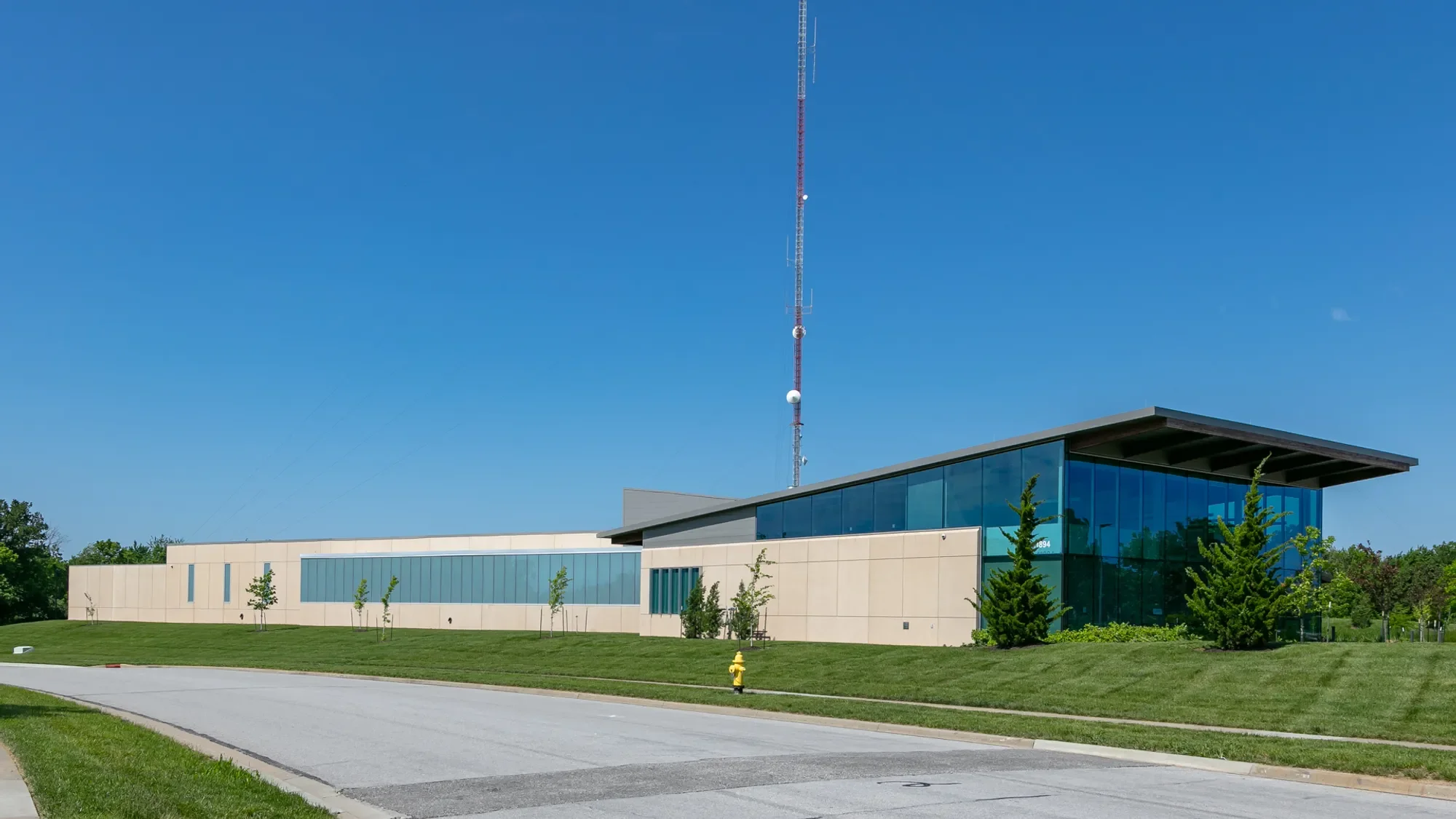 Modern building with glass windows and beige walls, surrounded by green grass and small trees, with a tall red-and-white communication tower against a clear blue sky.