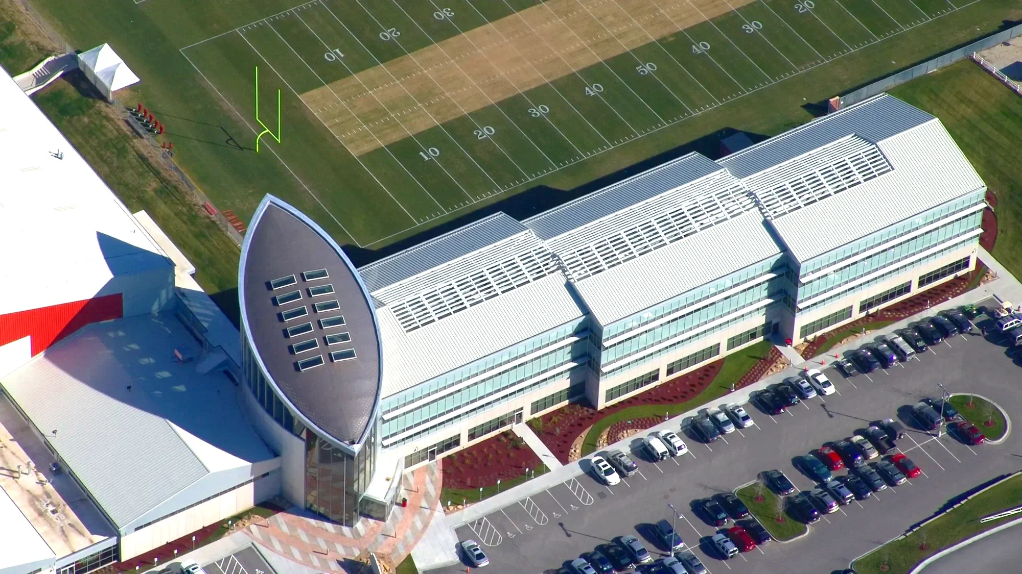 An aerial view of a modern building with a parking lot filled with cars in front of it. The building has a unique architectural design with large glass windows and a curved roof. There is a football field adjacent to the building, with marked yard lines and a field goal post visible.