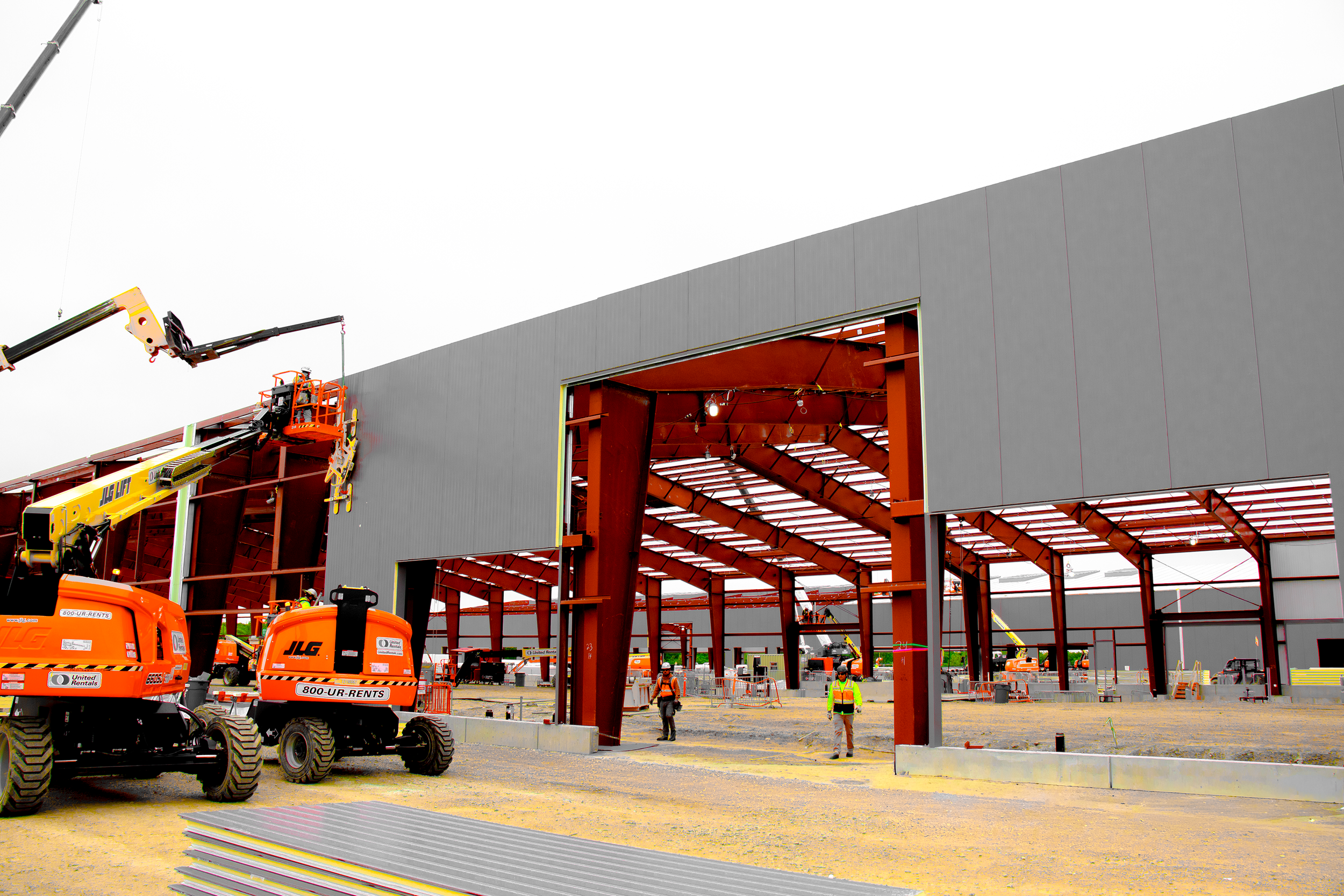 Construction site with workers and equipment building a large, gray warehouse with an open section showing internal red steel framework. Several orange lift machines and construction tools are visible in the foreground.