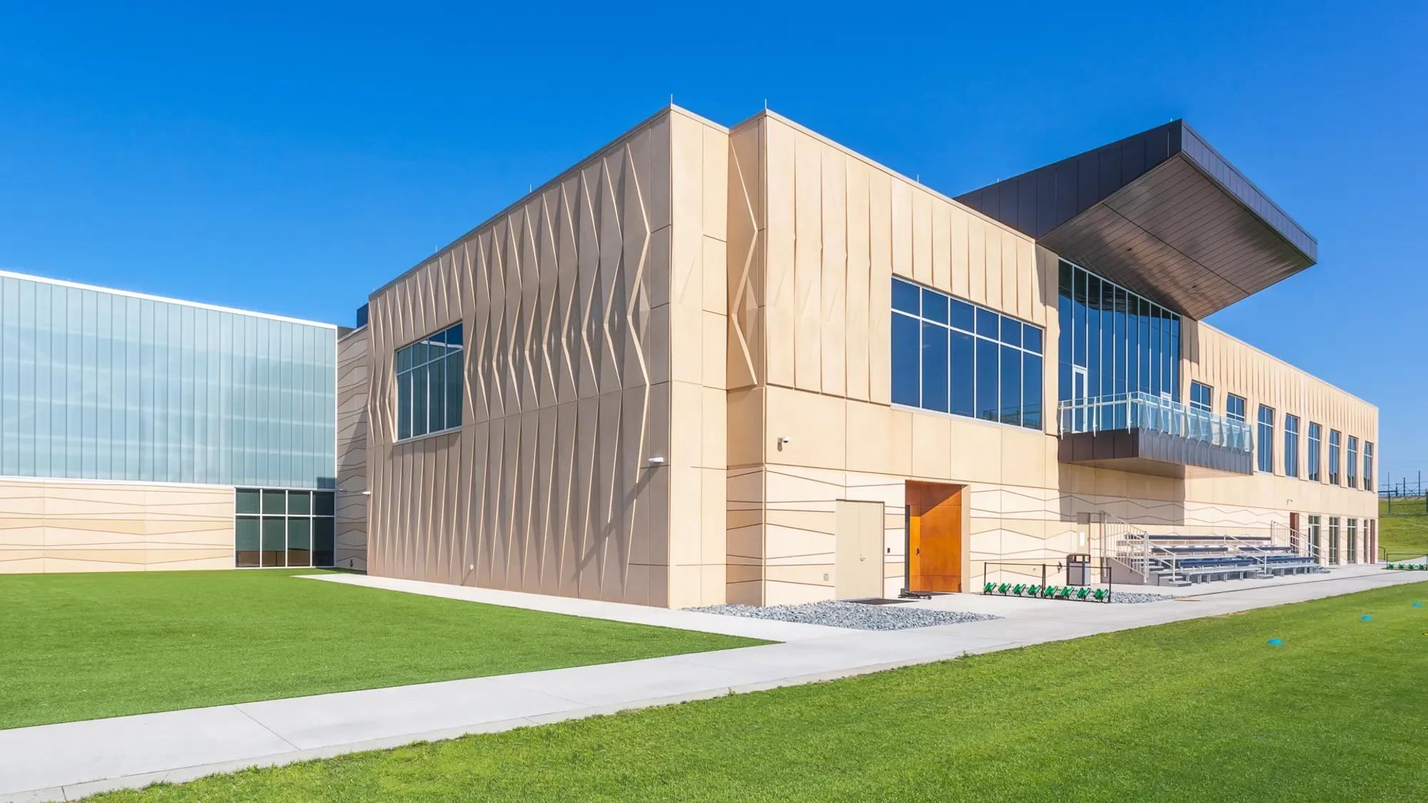 Modern building with beige and glass exterior, large windows, flat roof with overhang, green lawn in foreground, clear blue sky in background.