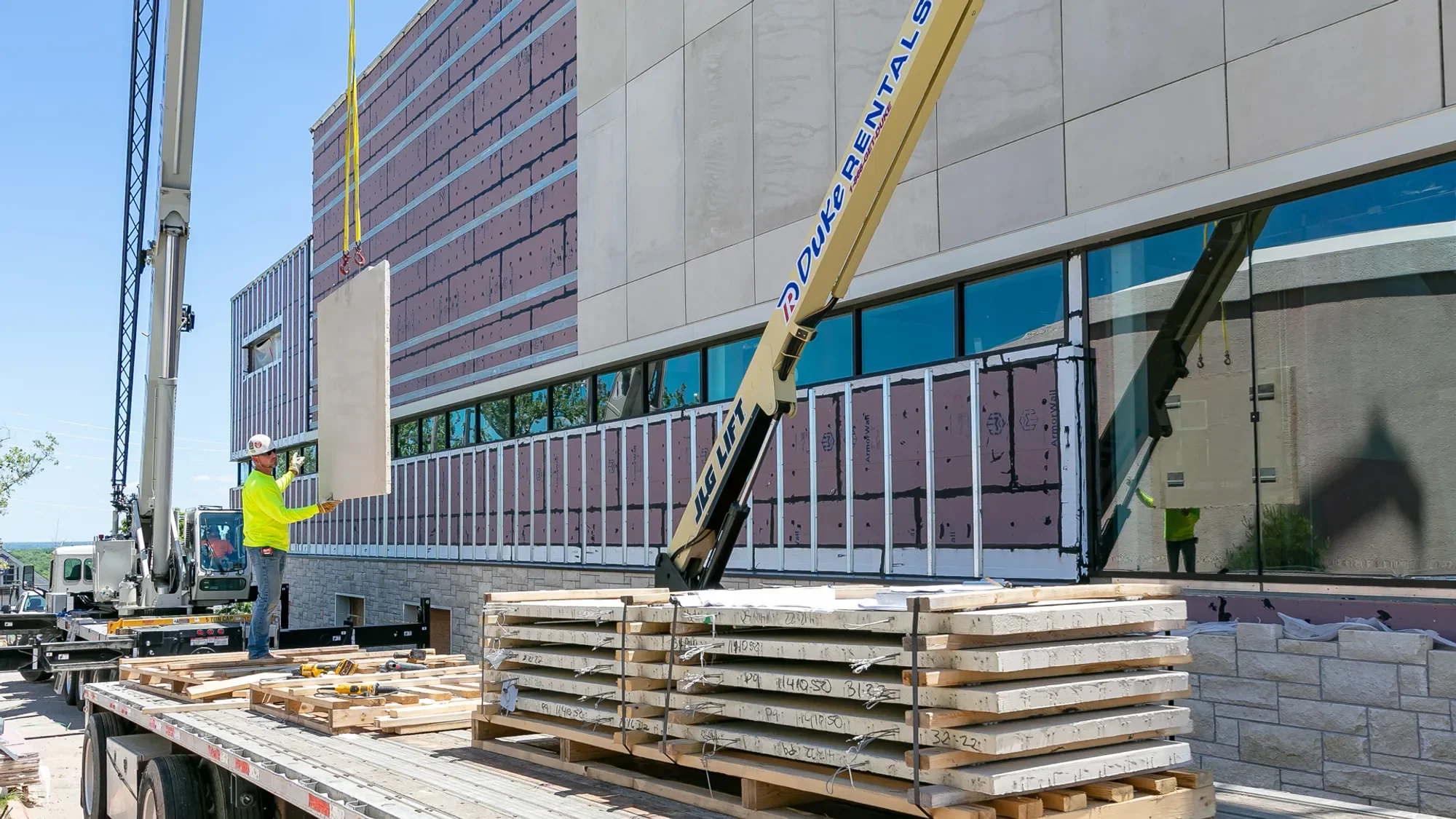 Construction workers install large concrete panels on the exterior of a modern building using a crane, with construction materials and tools in the foreground.